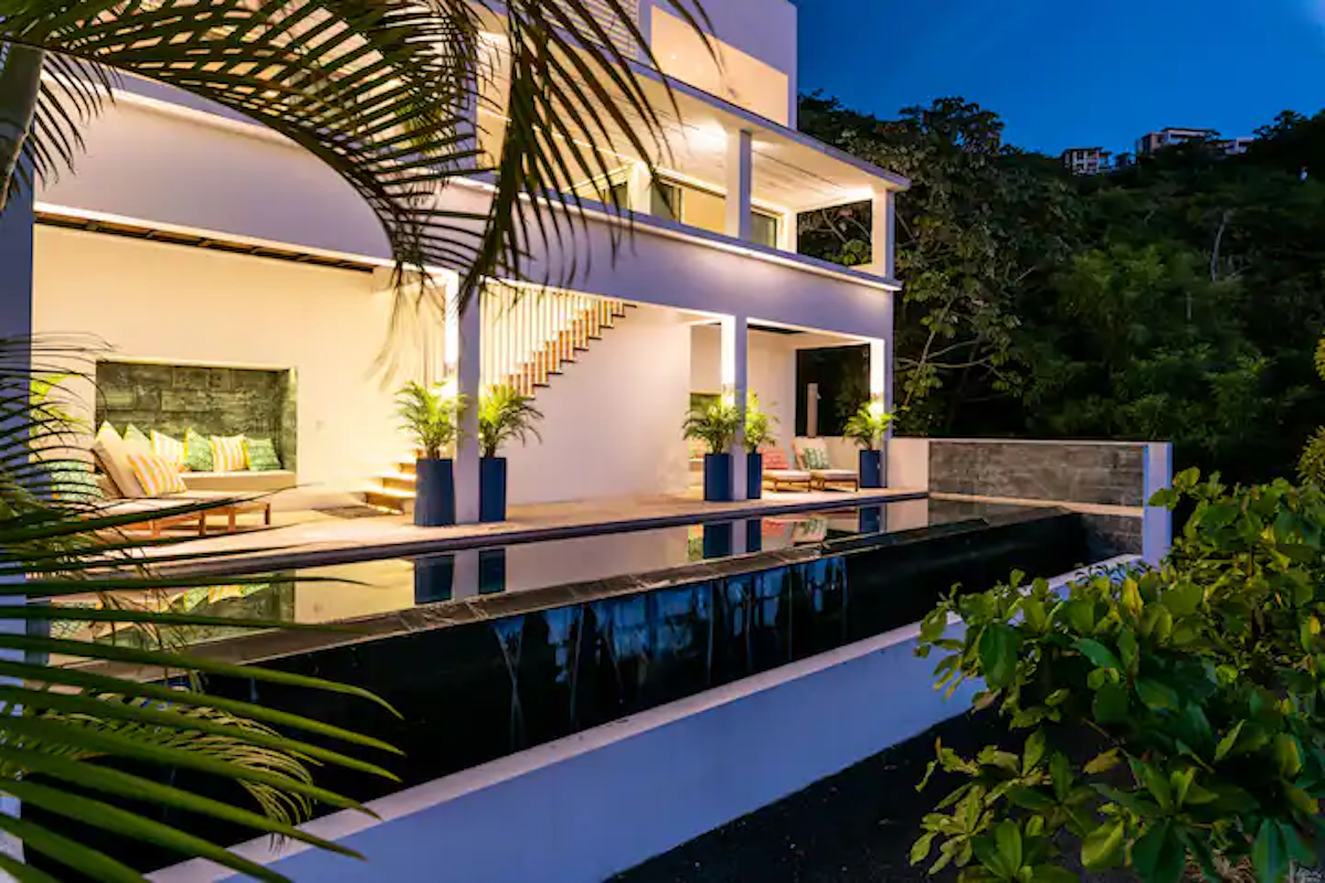 Maison blanche à deux étages avec une piscine rectangulaire sombre la nuit. Palmiers et verdure.