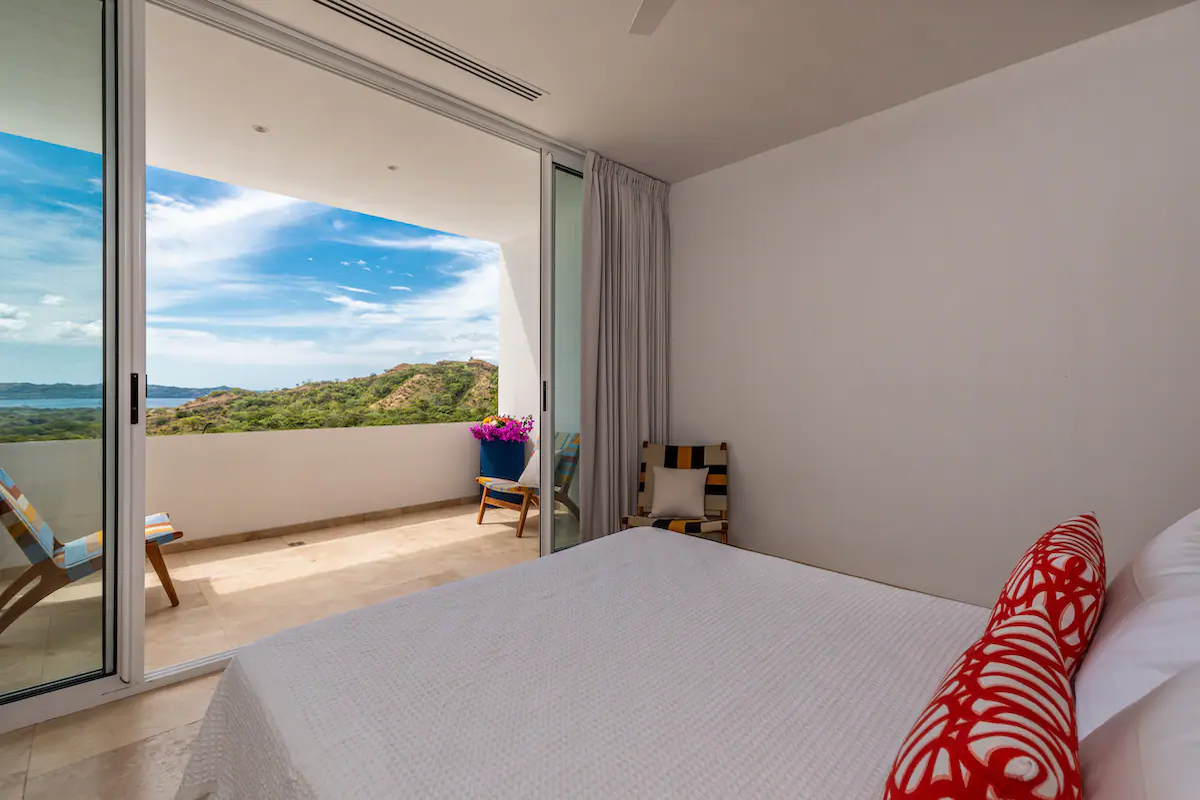 Chambre avec balcon donnant sur un paysage côtier ; lit blanc, porte-fenêtre coulissante.