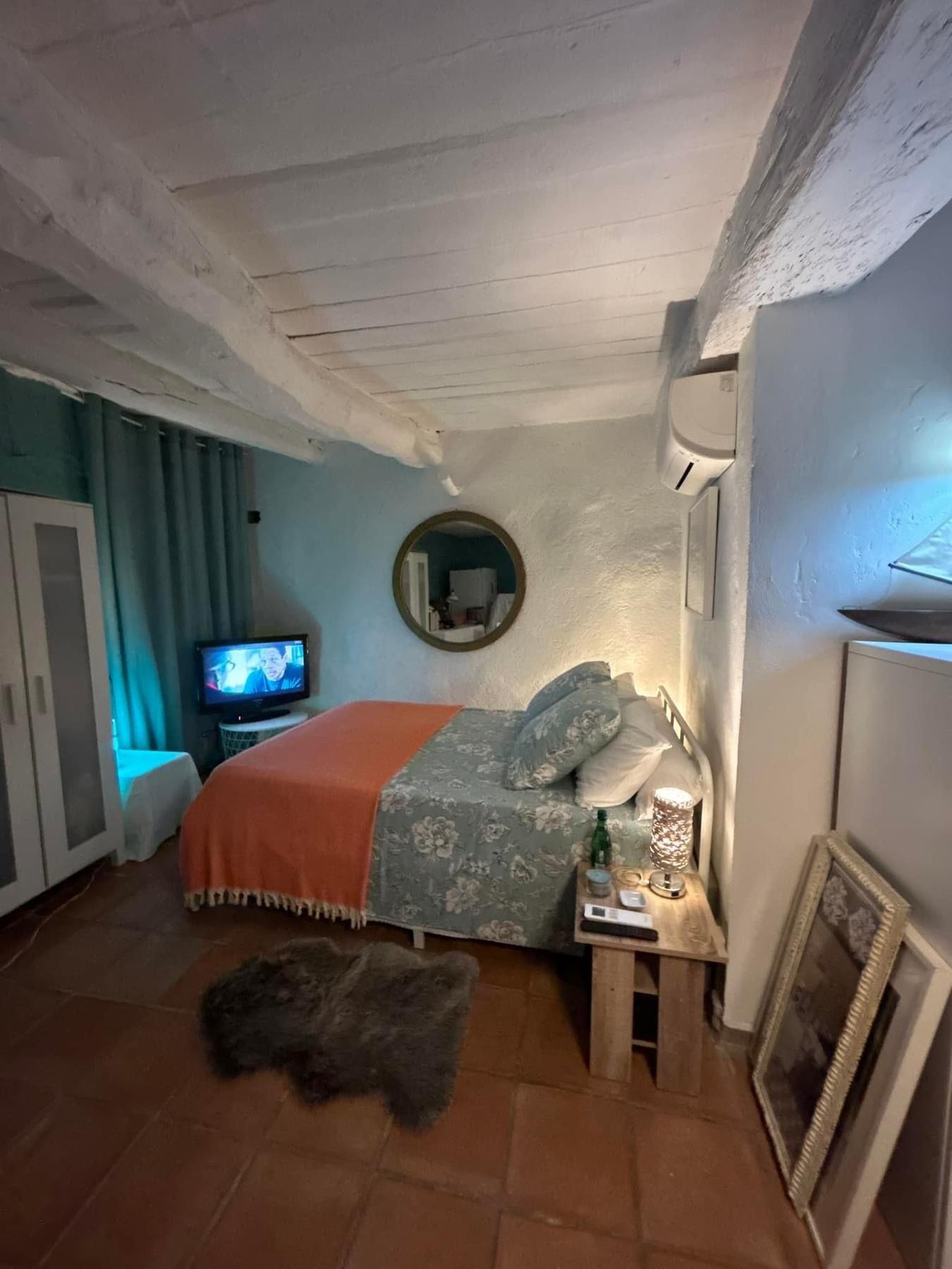 Chambre avec lit, tapis, télévision et miroir rond. Mur turquoise, poutres blanches au plafond et sol en terre cuite.