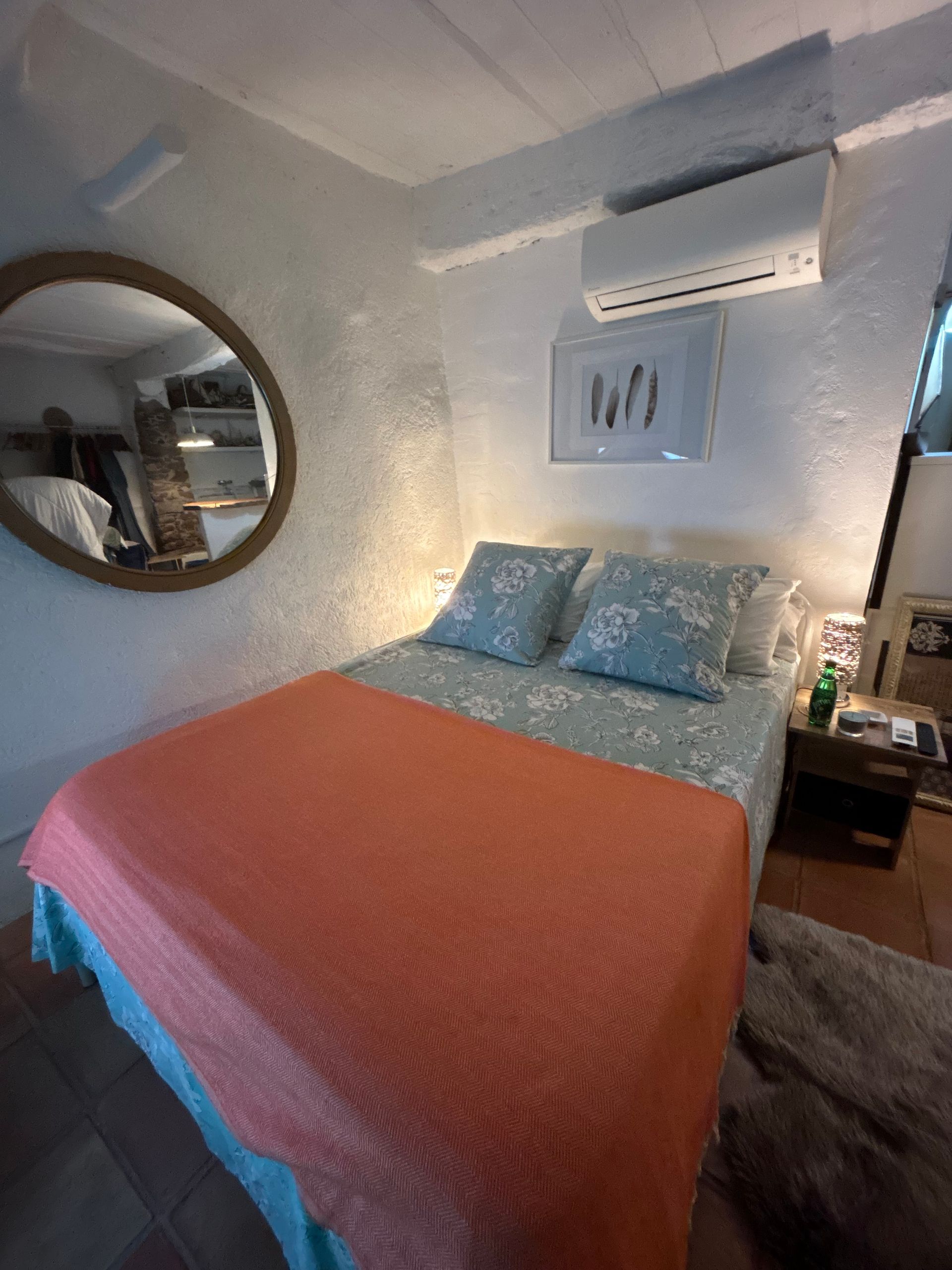 Chambre avec lit double, couverture corail, linge de lit à motifs floraux, miroir rond, murs blancs texturés.