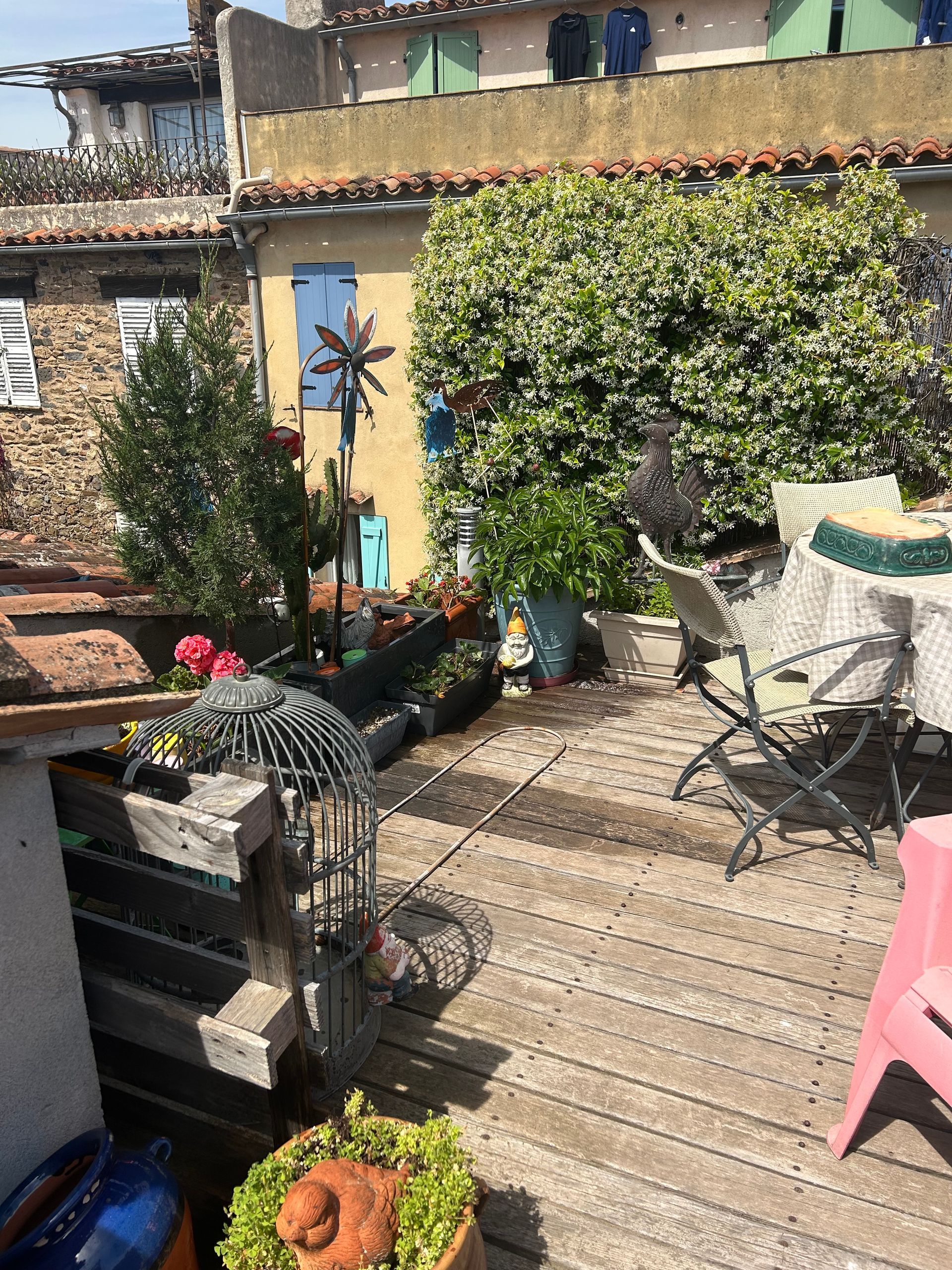 Terrasse sur le toit avec plancher en bois, plantes, table, chaise et girouette. Bâtiments en arrière-plan.