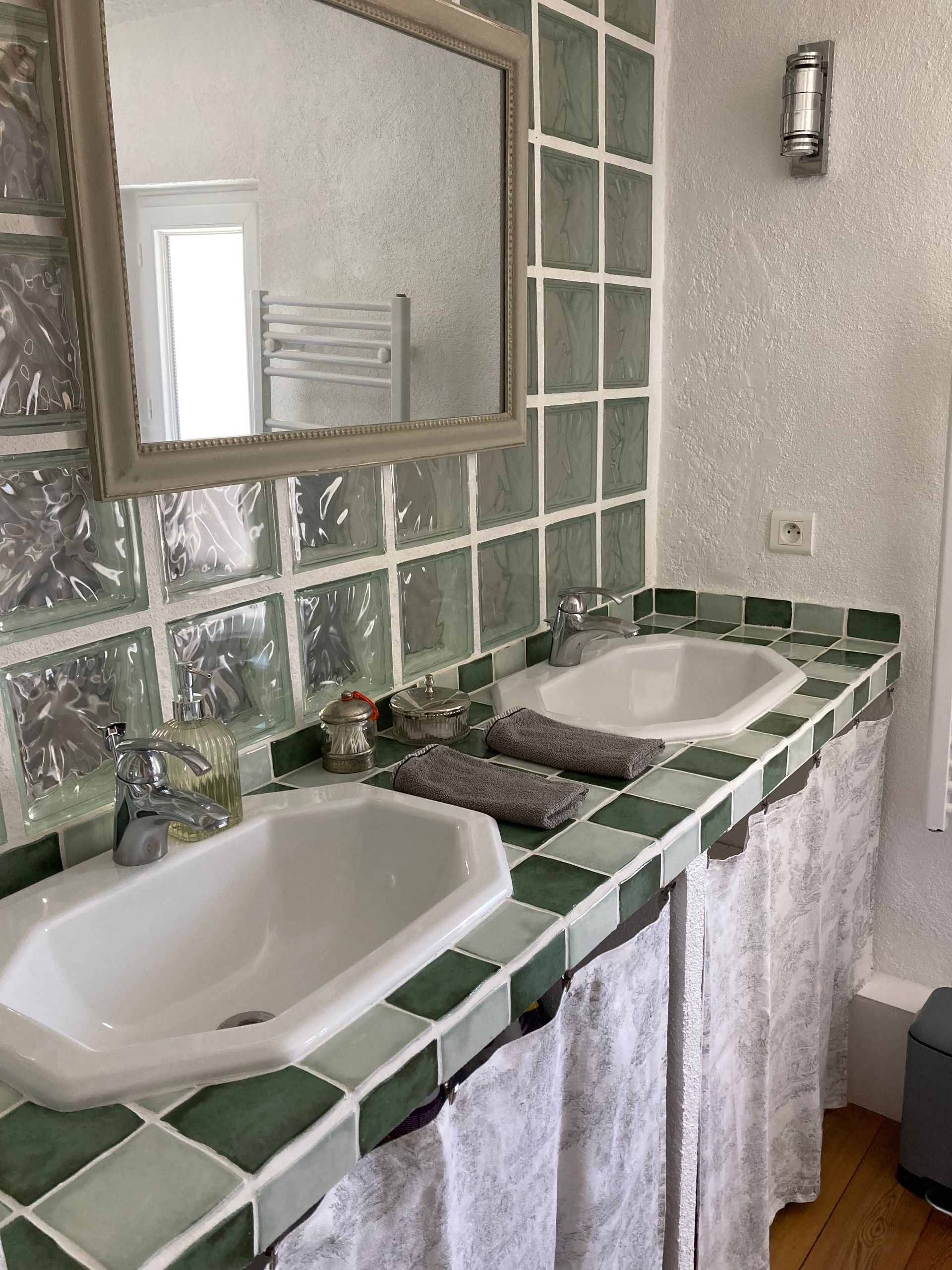 Salle de bains avec deux lavabos, un miroir et un mur carrelé de vert et de blanc. Lavabos, serviettes et rideaux blancs.