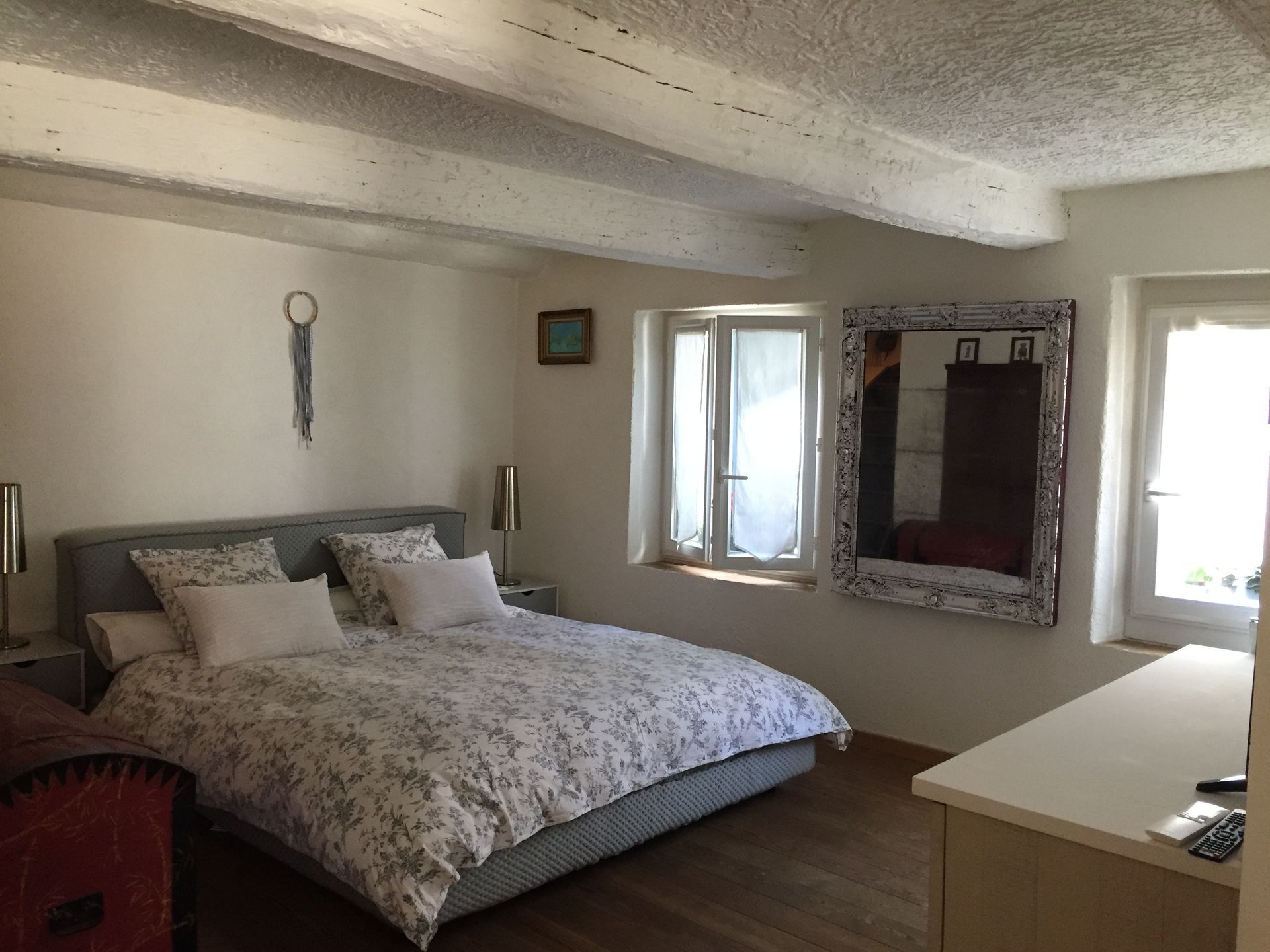 Chambre avec lit, commode, fenêtres et grand miroir. Murs blancs et poutres apparentes.
