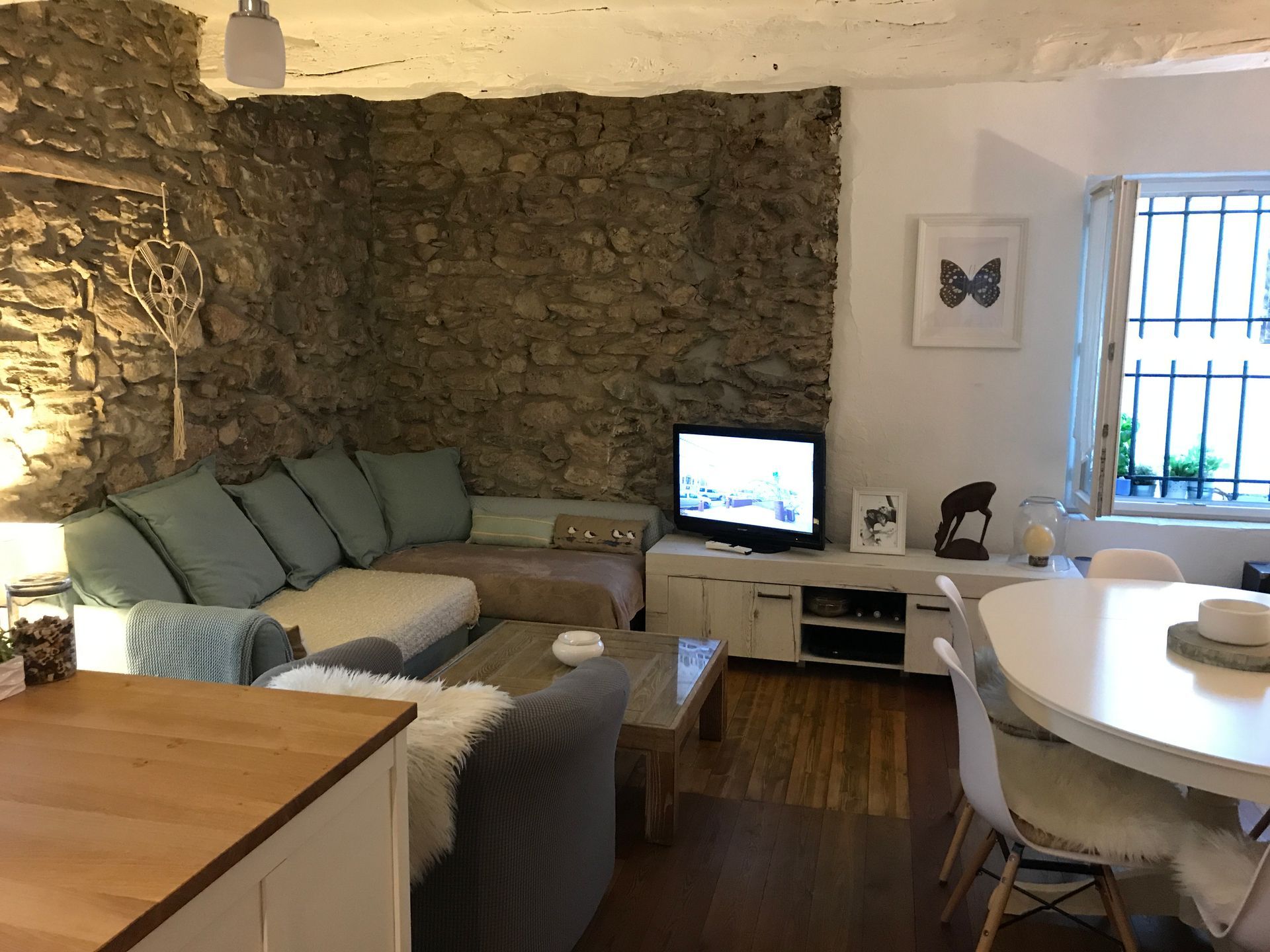Salon avec mur en pierre, canapé d'angle, télévision et table à manger.