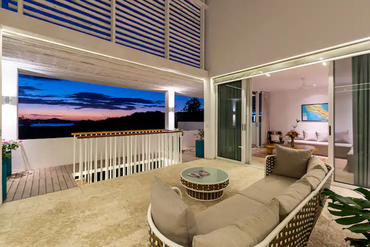 Balcon avec canapé beige, vue sur le ciel du soir. Une rambarde blanche et des portes coulissantes mènent à l'intérieur.