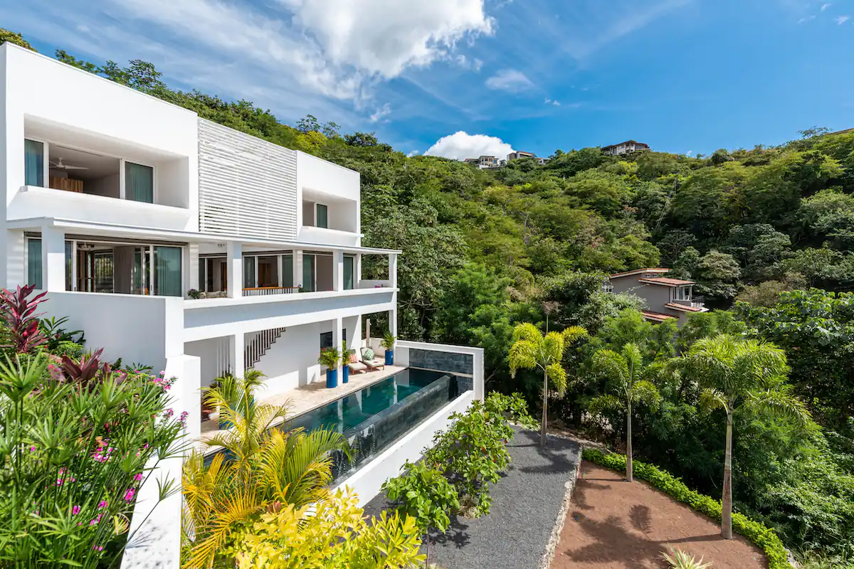 Villa blanche moderne avec piscine et végétation luxuriante sous un ciel d'un bleu éclatant.