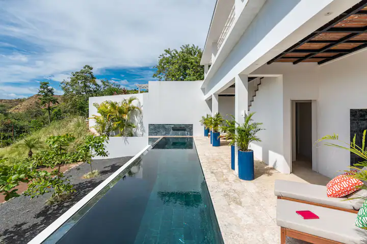 Piscine à débordement jouxtant un bâtiment blanc orné de plantes en pot bleues et offrant une vue sur les collines verdoyantes.