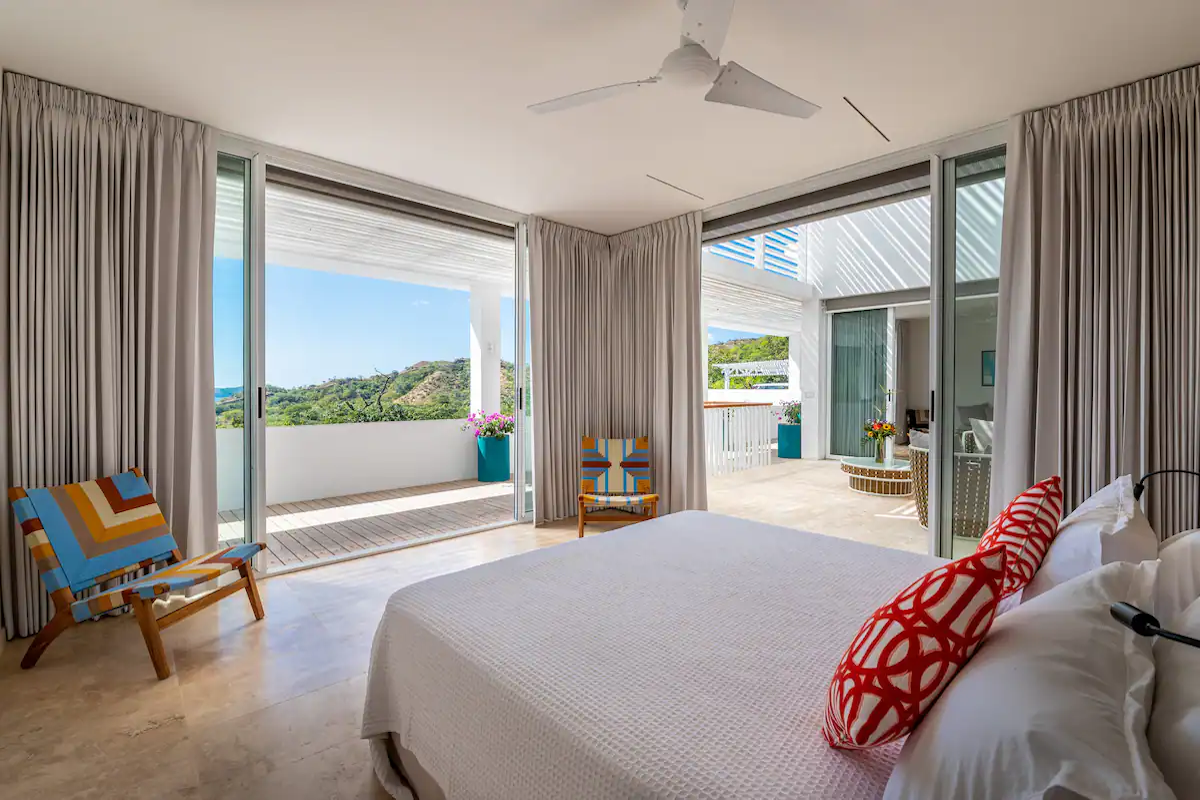 Chambre avec portes ouvertes donnant sur un balcon avec vue sur le paysage ; touches de crème et de turquoise.