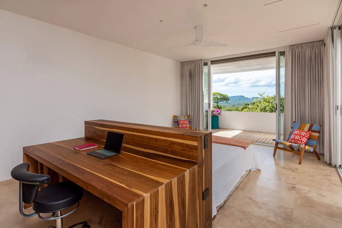 Chambre avec bureau et balcon donnant sur un paysage luxuriant ; portes coulissantes ouvertes, murs blancs.