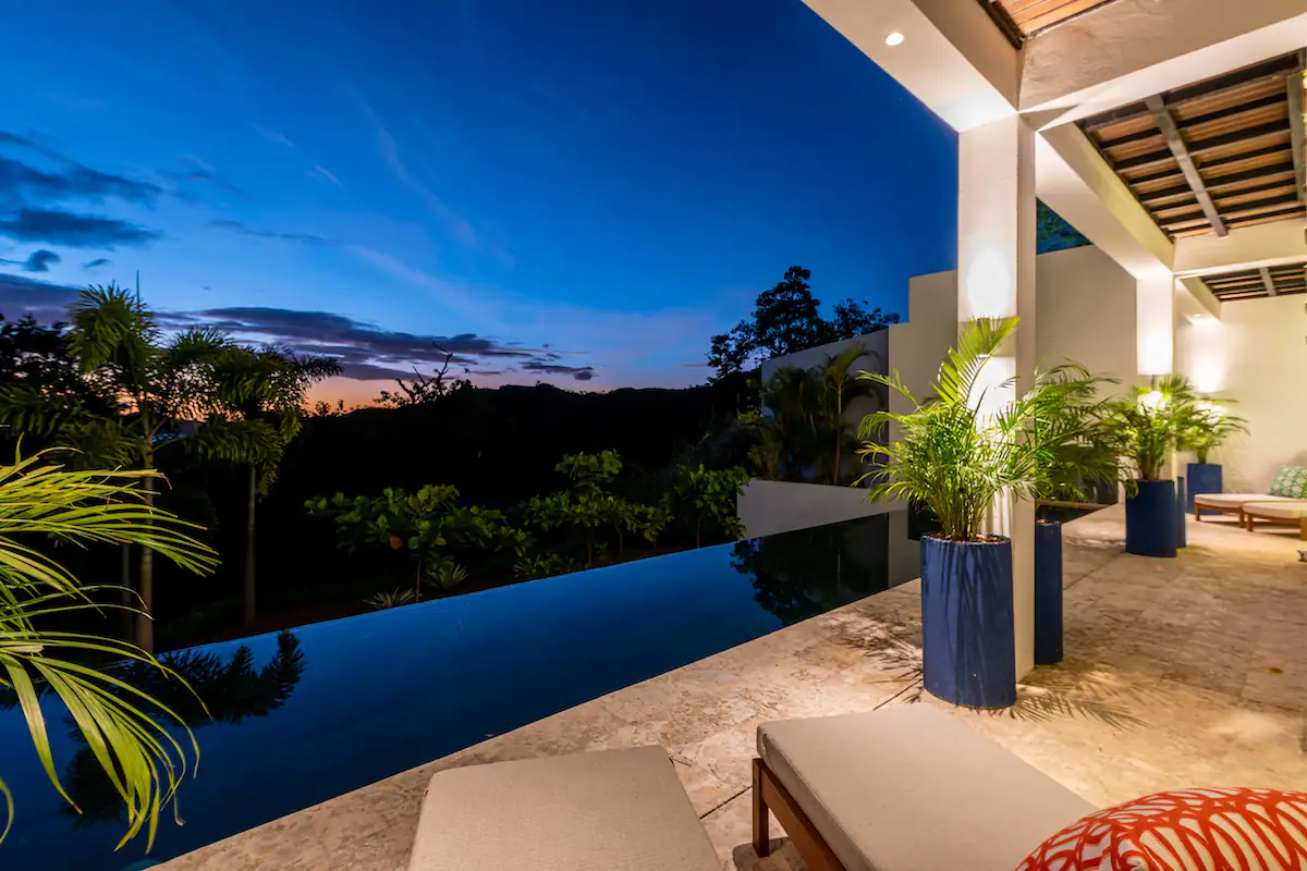 Piscine à débordement donnant sur un paysage tropical au crépuscule. Chaises longues et plantes en pot à proximité.
