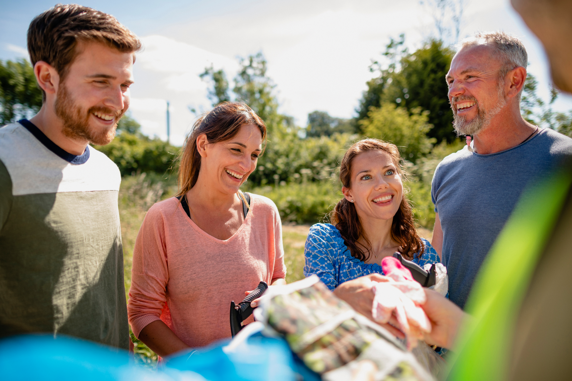 A group of four adults smiling and talking outdoors in a bright, grassy setting, with one person holding a blue bag.