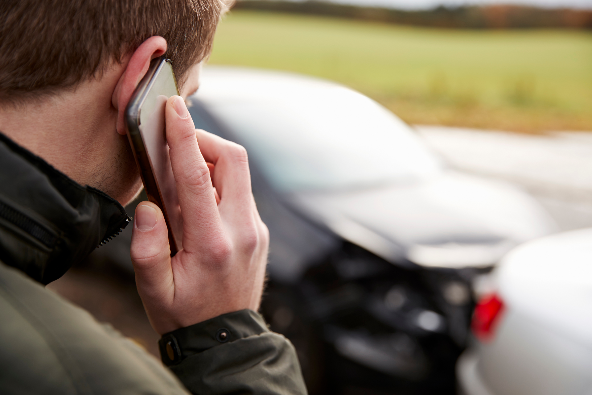 A person makes a phone call with a damaged car in the background after an accident.