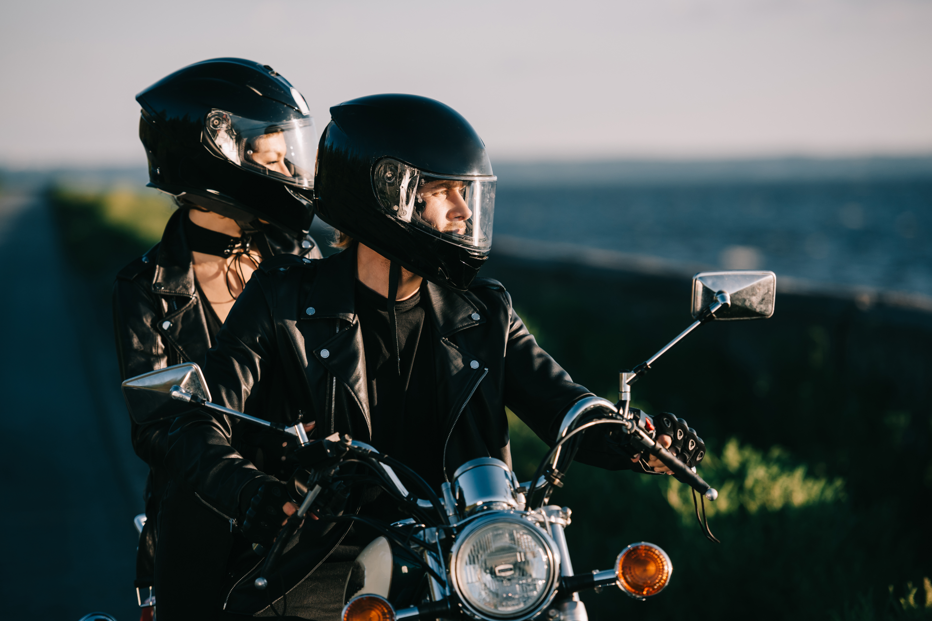 Two people wearing black helmets and leather jackets riding a motorcycle along a road near a body of water.