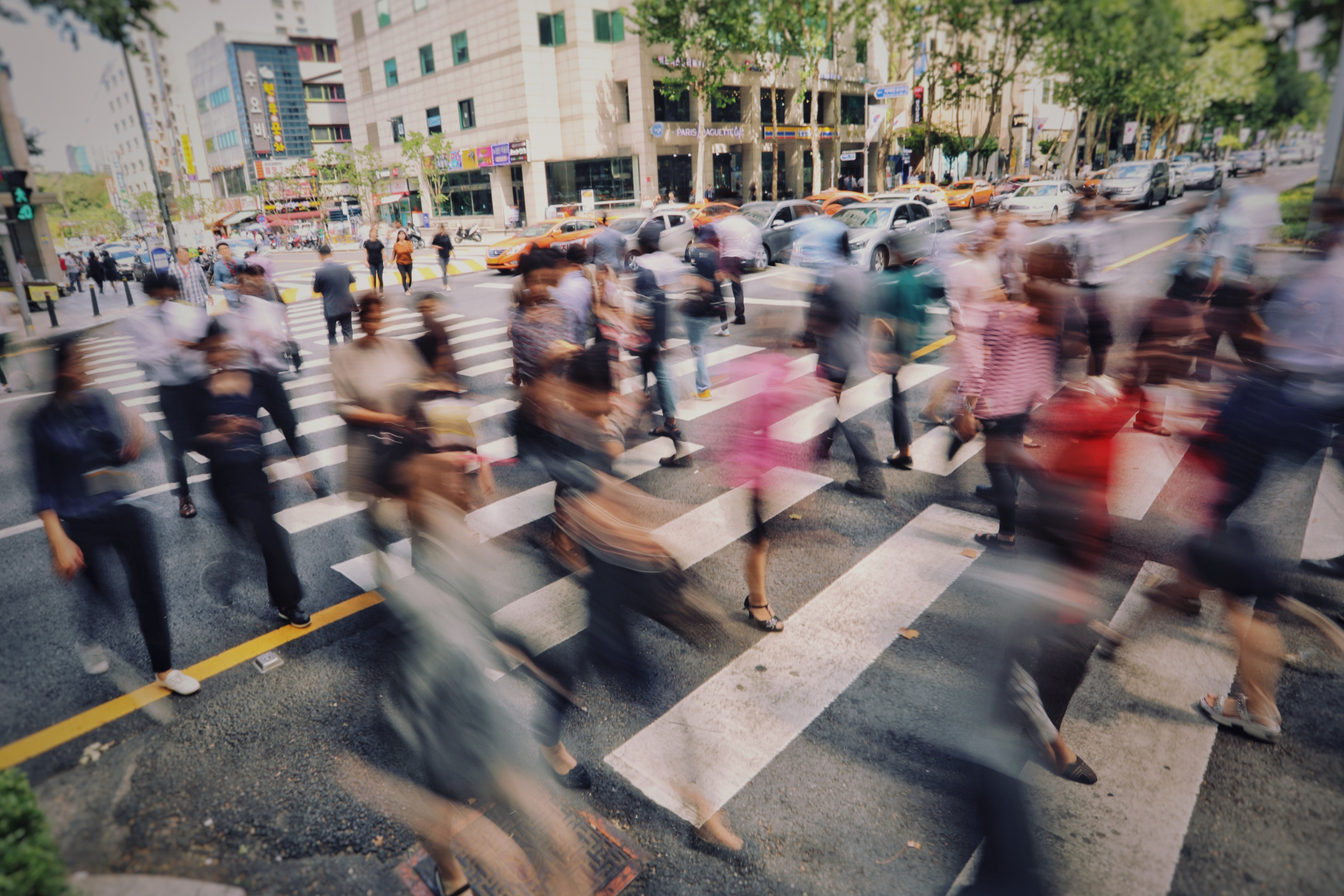 A motion-blurred view of pedestrians walking across a city crosswalk on a busy street during the day.