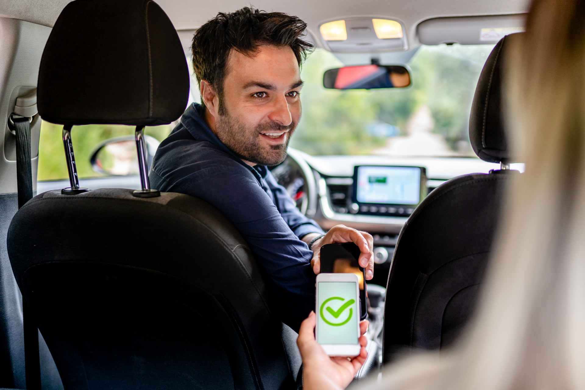A person in the driver's seat of a car shows a smartphone screen with a green checkmark to a passenger in the back.