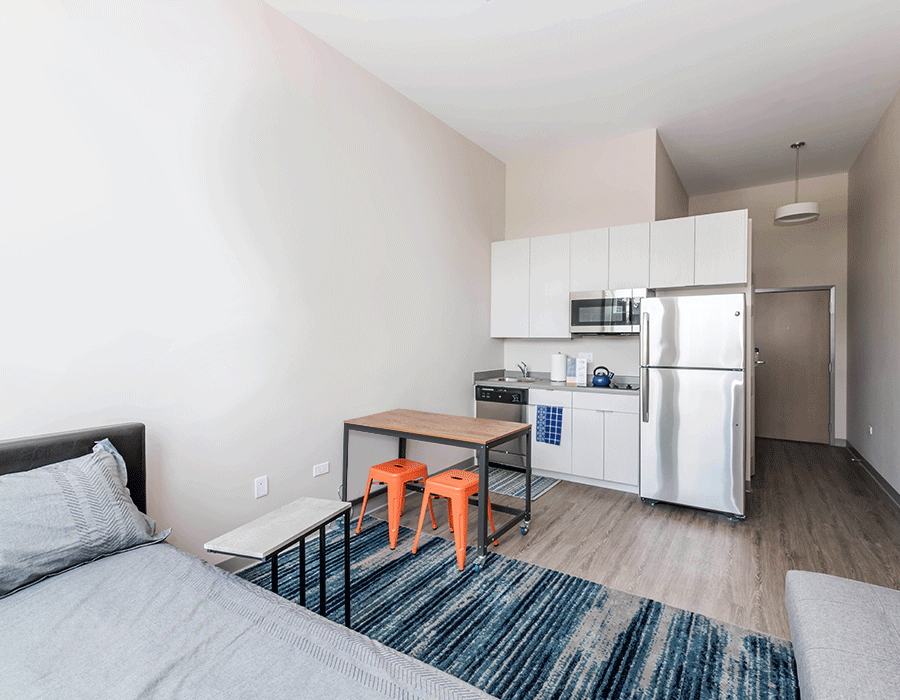 A bedroom with a bed , table , stools and a kitchen.