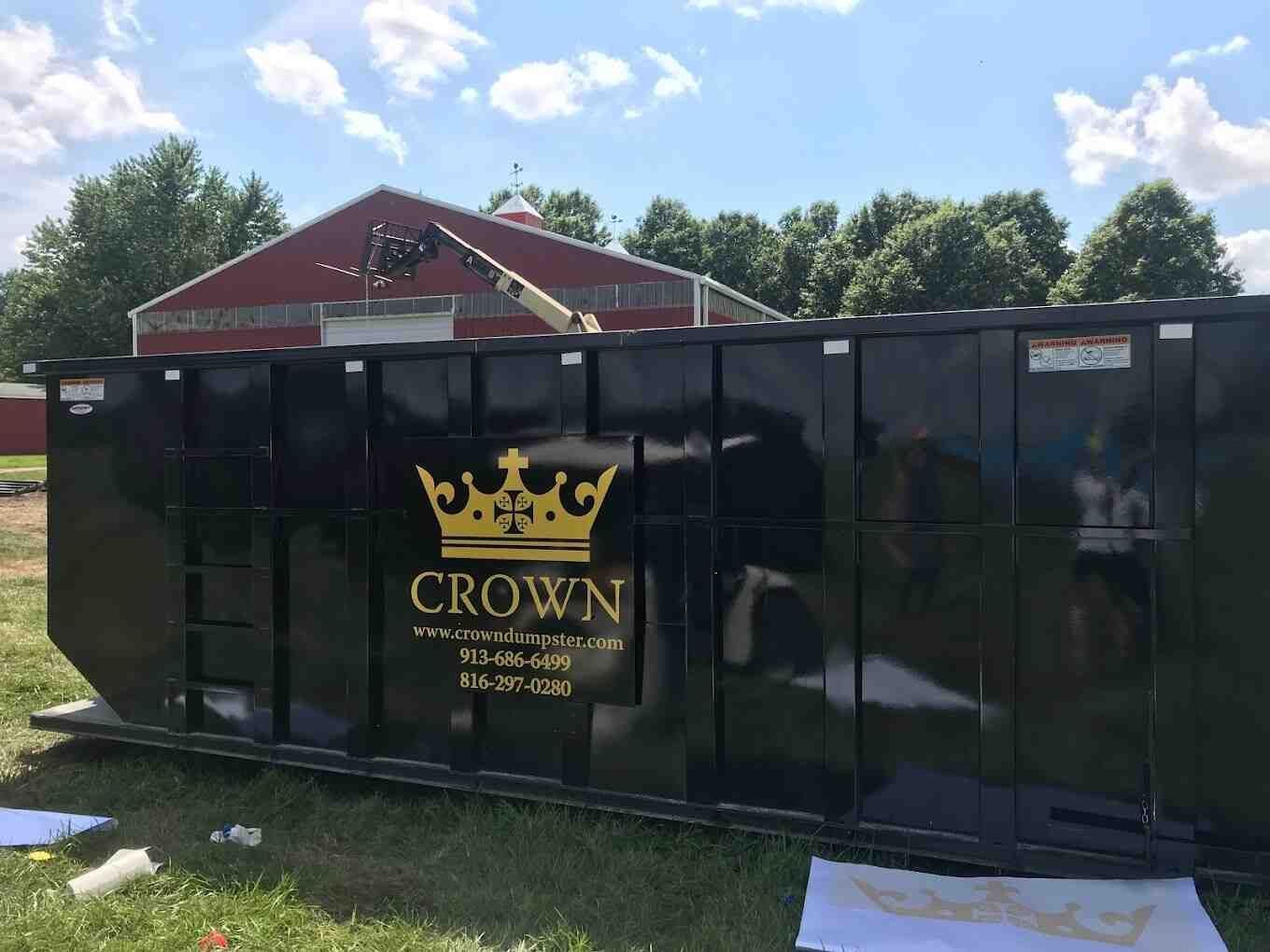 Black CROWN dumpster in a grassy yard with a red barn and trees in the background