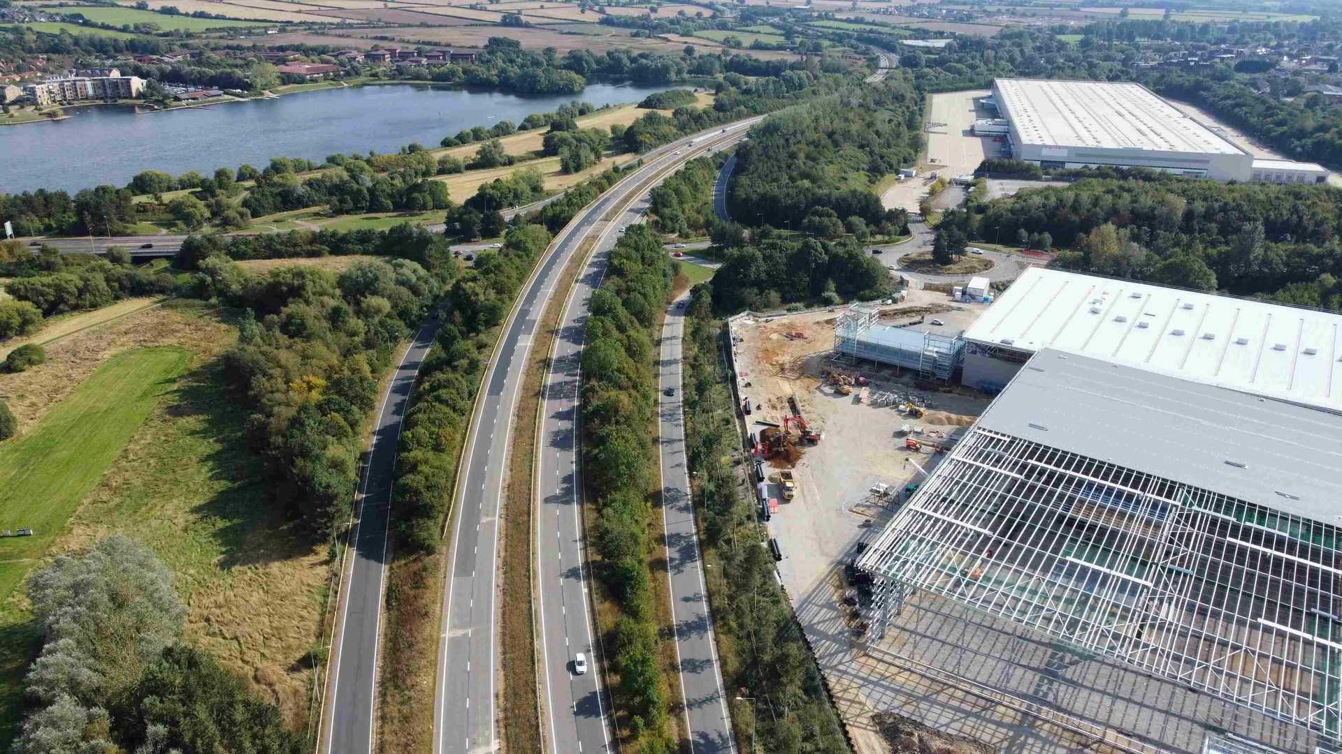 Aerial view of highway with several lanes, lined with trees, next to a lake and large industrial buildings.