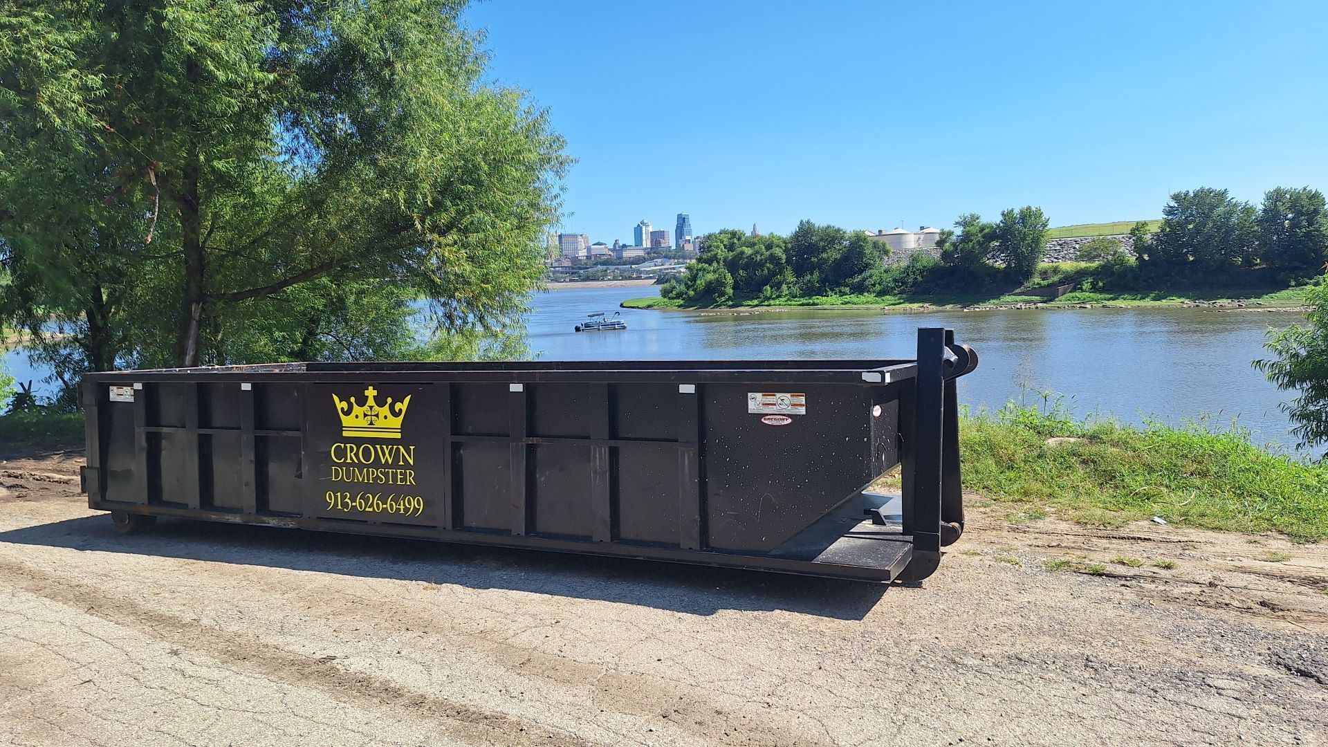 Black dumpster by a riverside path with trees and a city skyline in the distance
