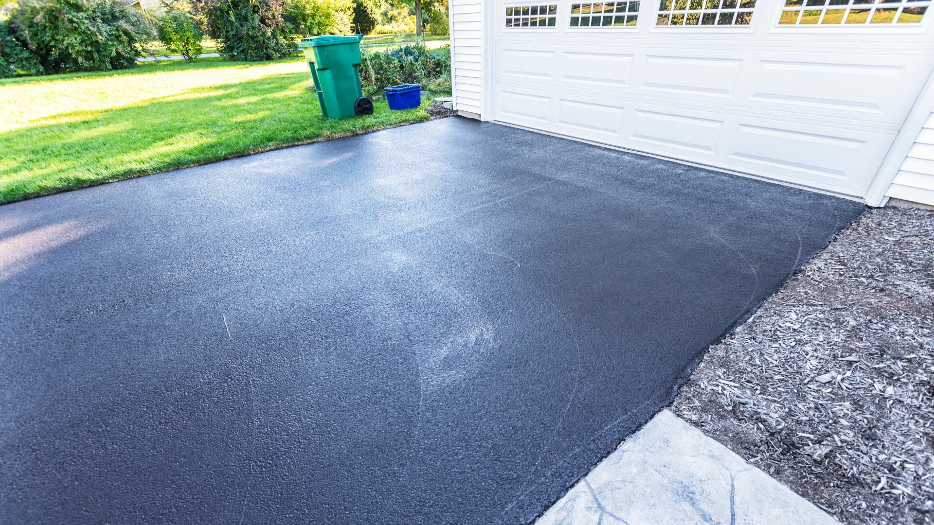 Newly sealed black asphalt driveway next to a white garage and a green lawn.