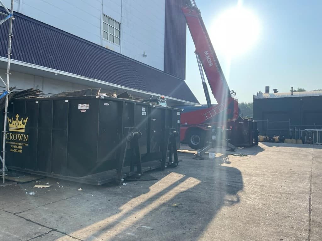 Black dumpster near a building; a red crane is extended towards it under the bright sun.