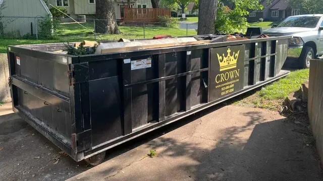 Black Crown dumpster on a residential driveway, filled with debris, sunny day.