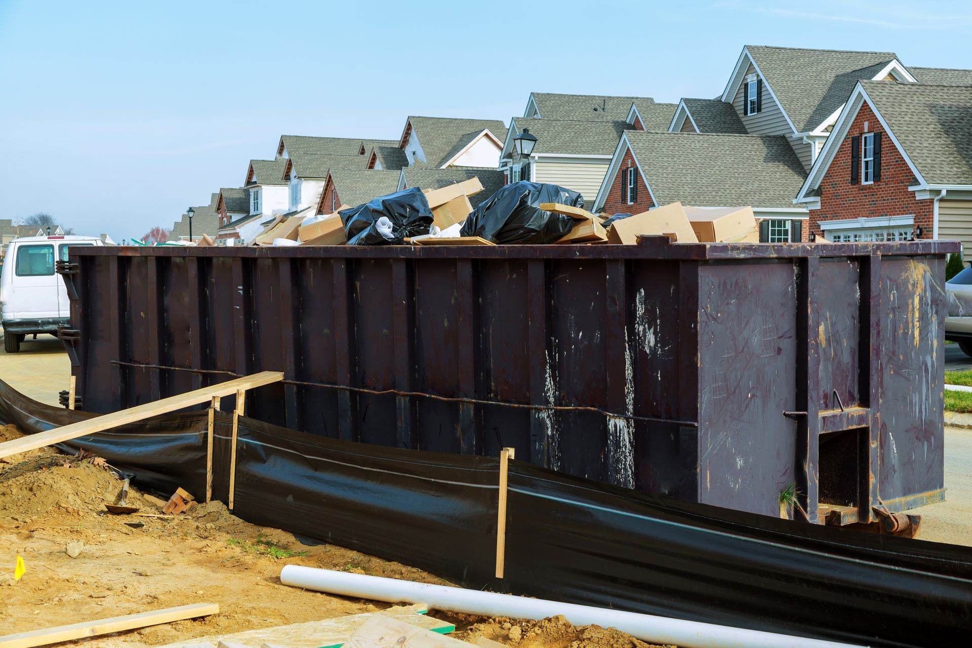A large, dark brown dumpster filled with construction debris sits in a residential neighborhood near a fenced construction site.
