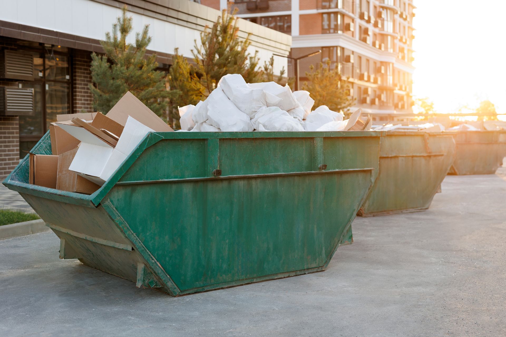Green construction dumpster filled with cardboard and trash on a city street at sunset