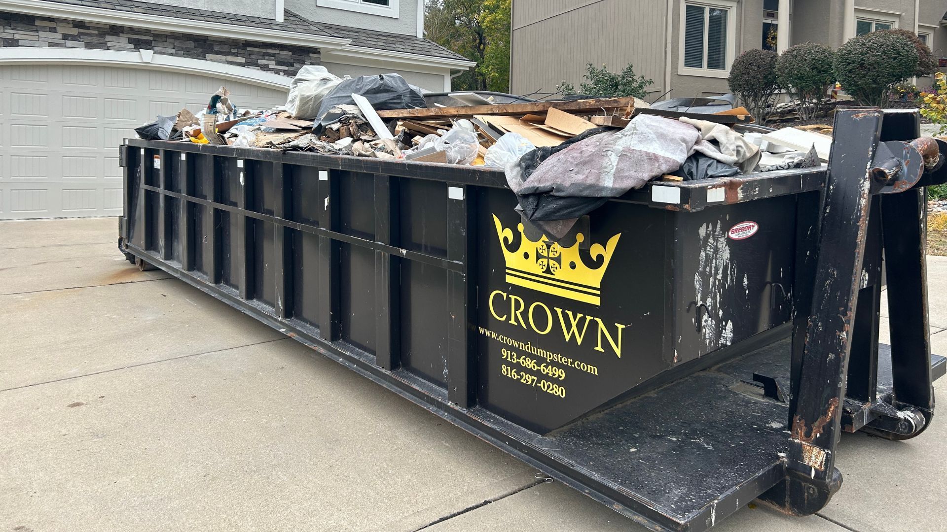 Black dumpster overflowing with debris parked on a driveway near a house.
