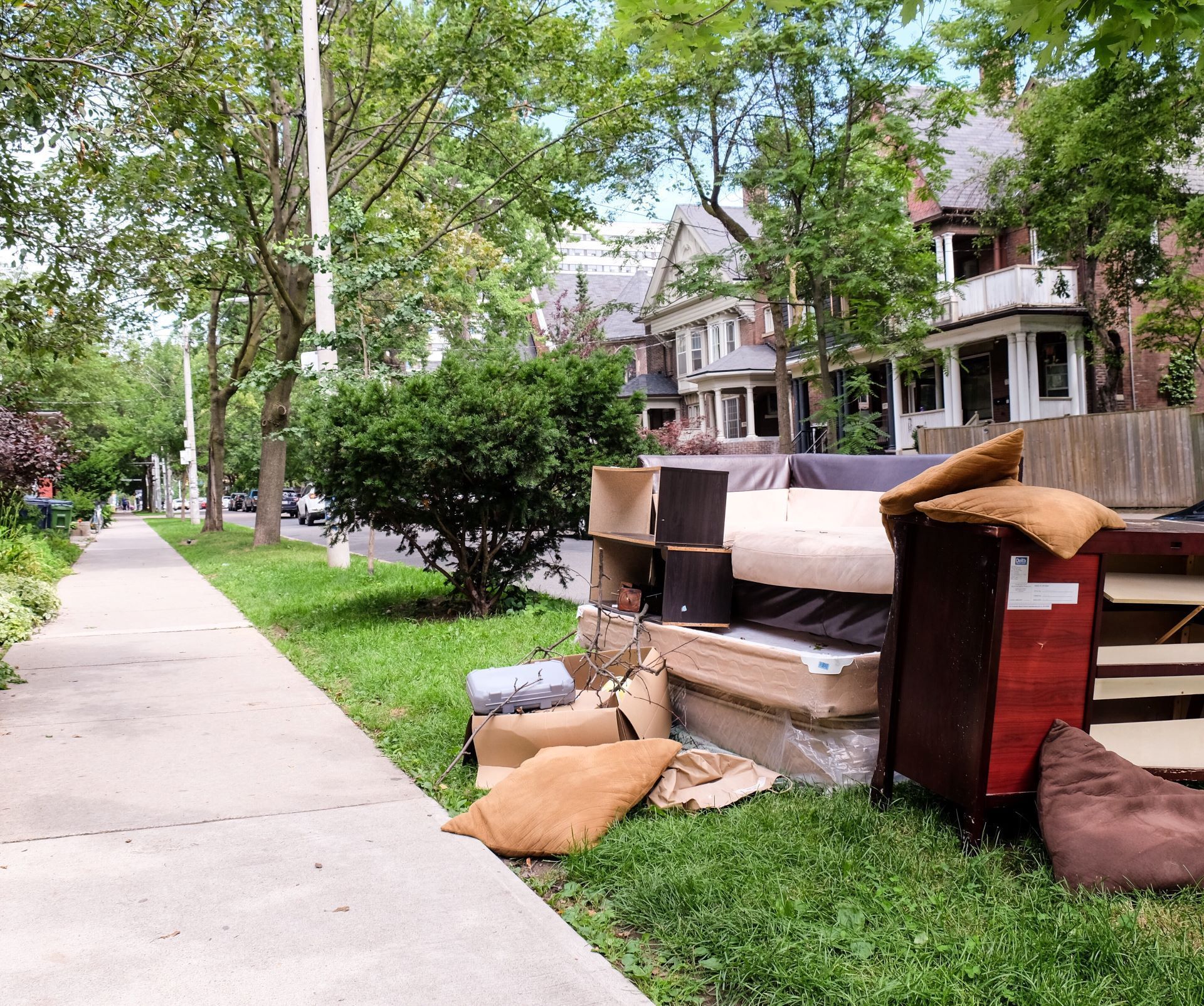 Sidewalk with discarded furniture piled on the grass near street. Houses and trees line the road.