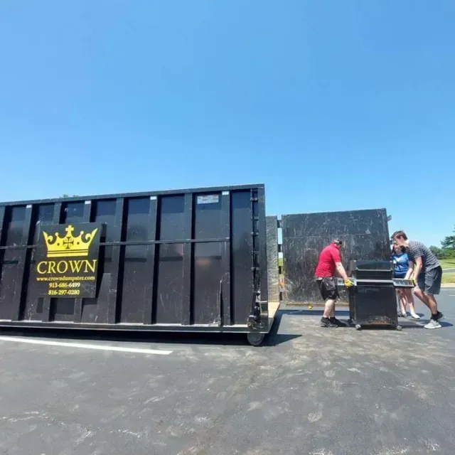 Two people loading a black grill into a large black dumpster under a clear blue sky.