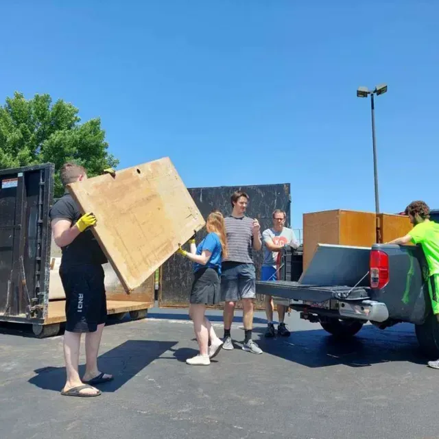 People loading a large wooden board into a truck bed with a dumpster nearby under a clear blue sky.