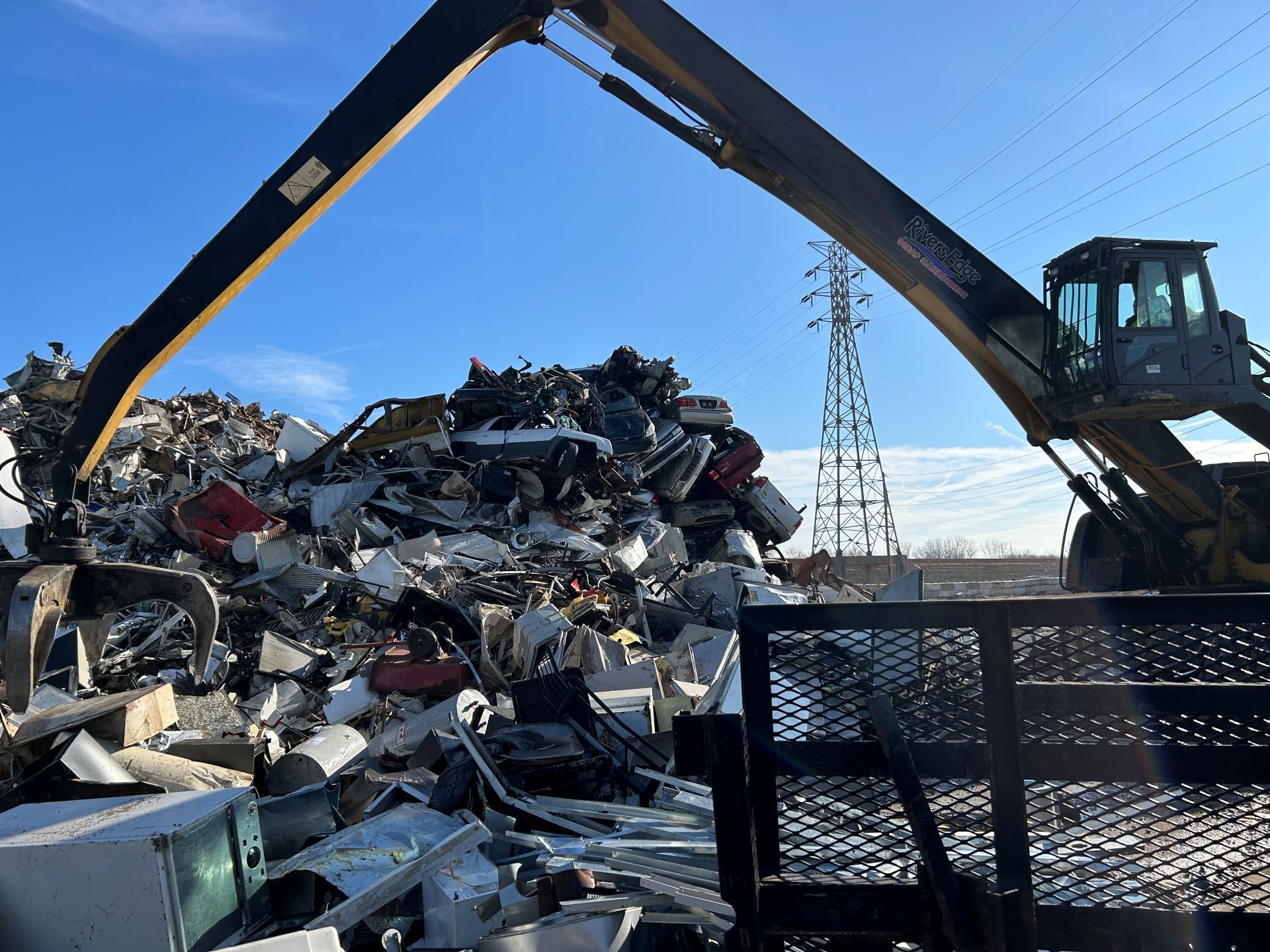A large excavator loading scrap metal onto a flatbed truck at a salvage yard under a blue sky.