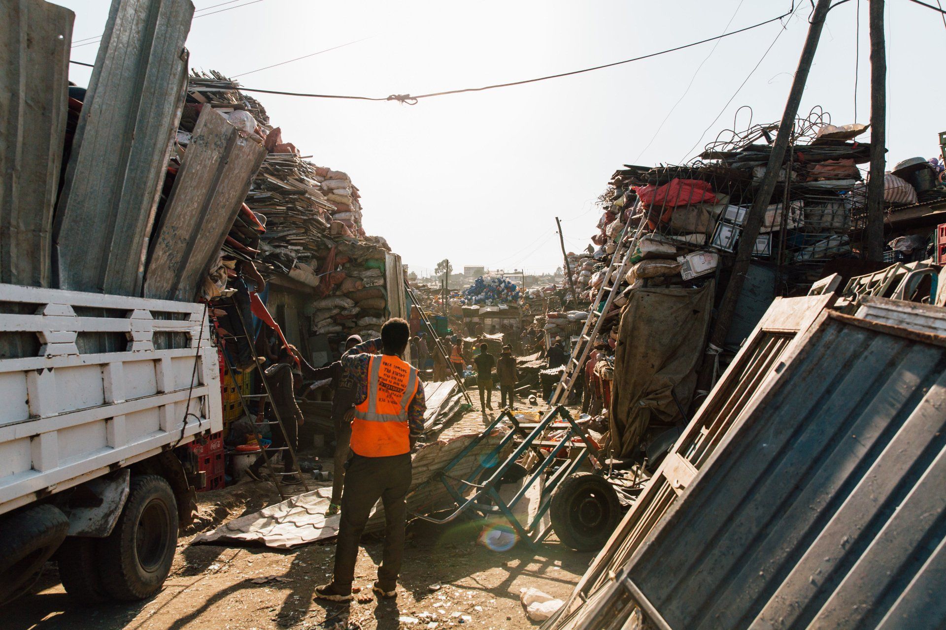 Man in orange vest in a cluttered junkyard loading a truck with scrap metal; sunny day.
