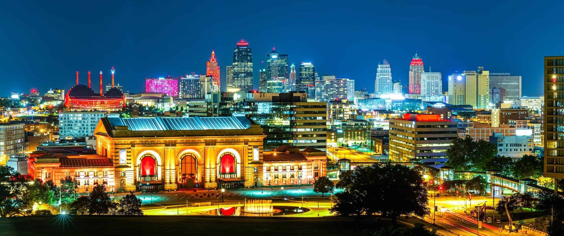 Night view of Kansas City skyline with lit-up buildings and structures. Blue sky above, yellow and red lights below.