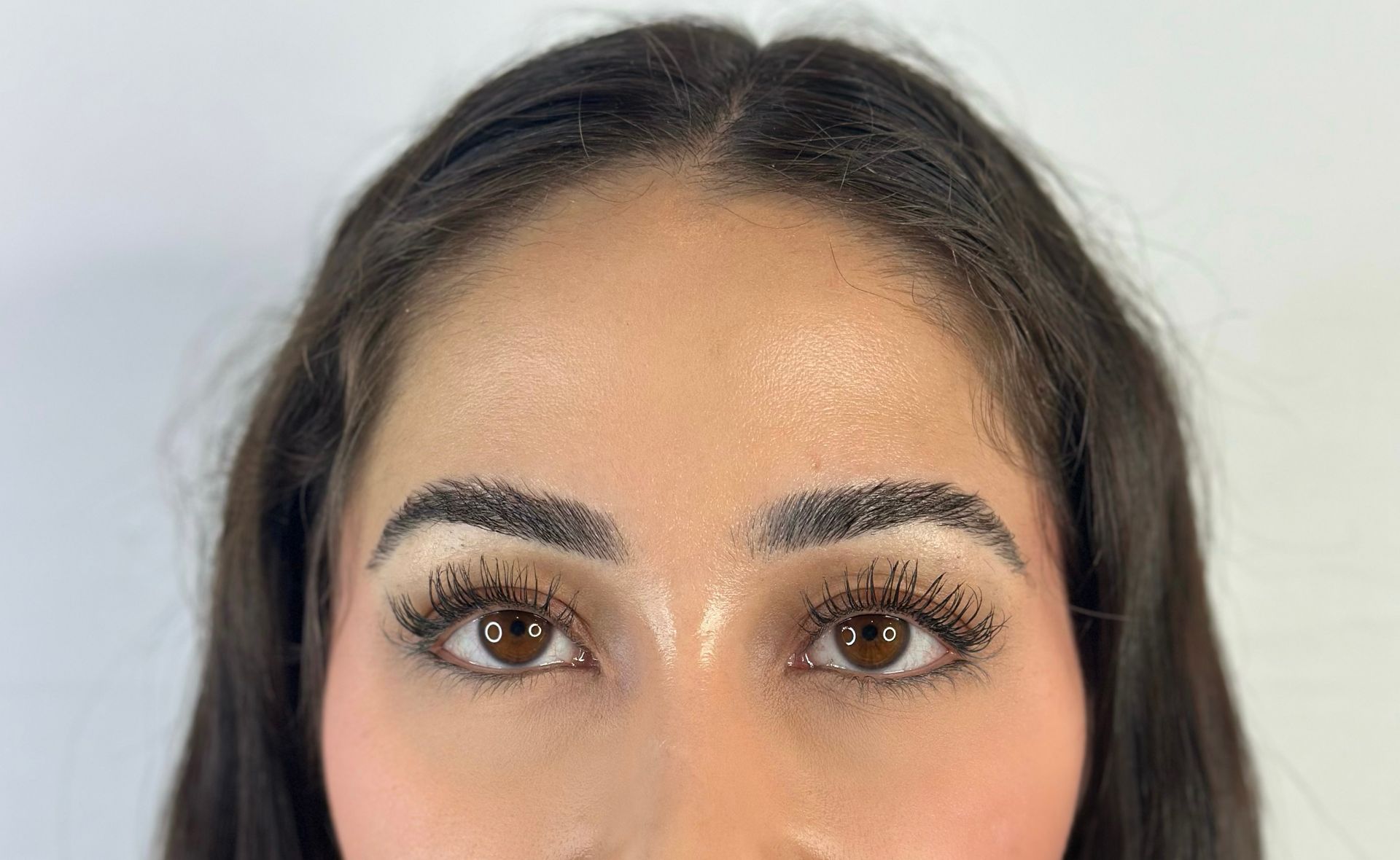 Close-up of a woman's face. Brown eyes, long lashes, filled eyebrows, and dark hair, against a light background.