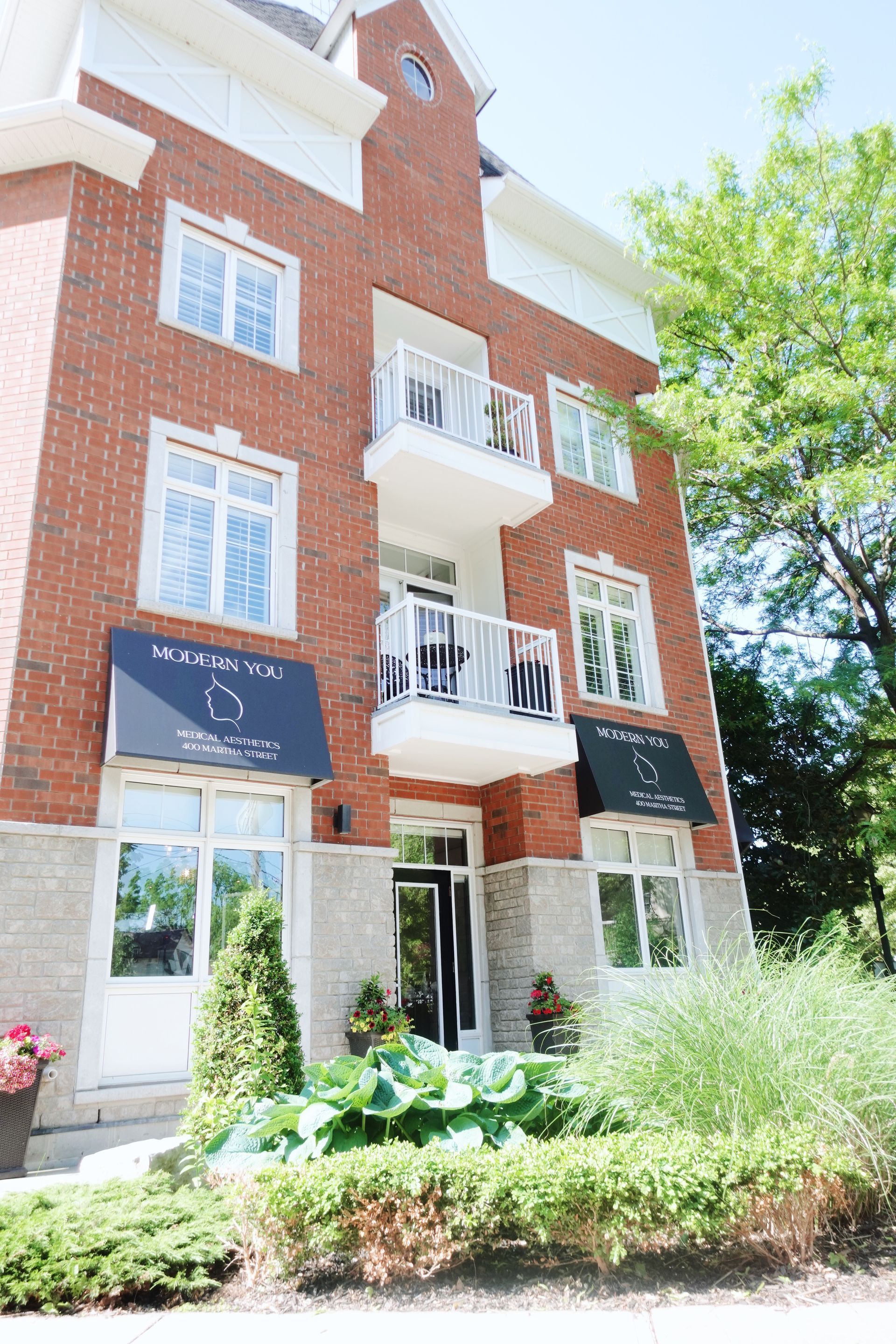 Brick building with balconies and black awnings over windows, lush greenery in front.