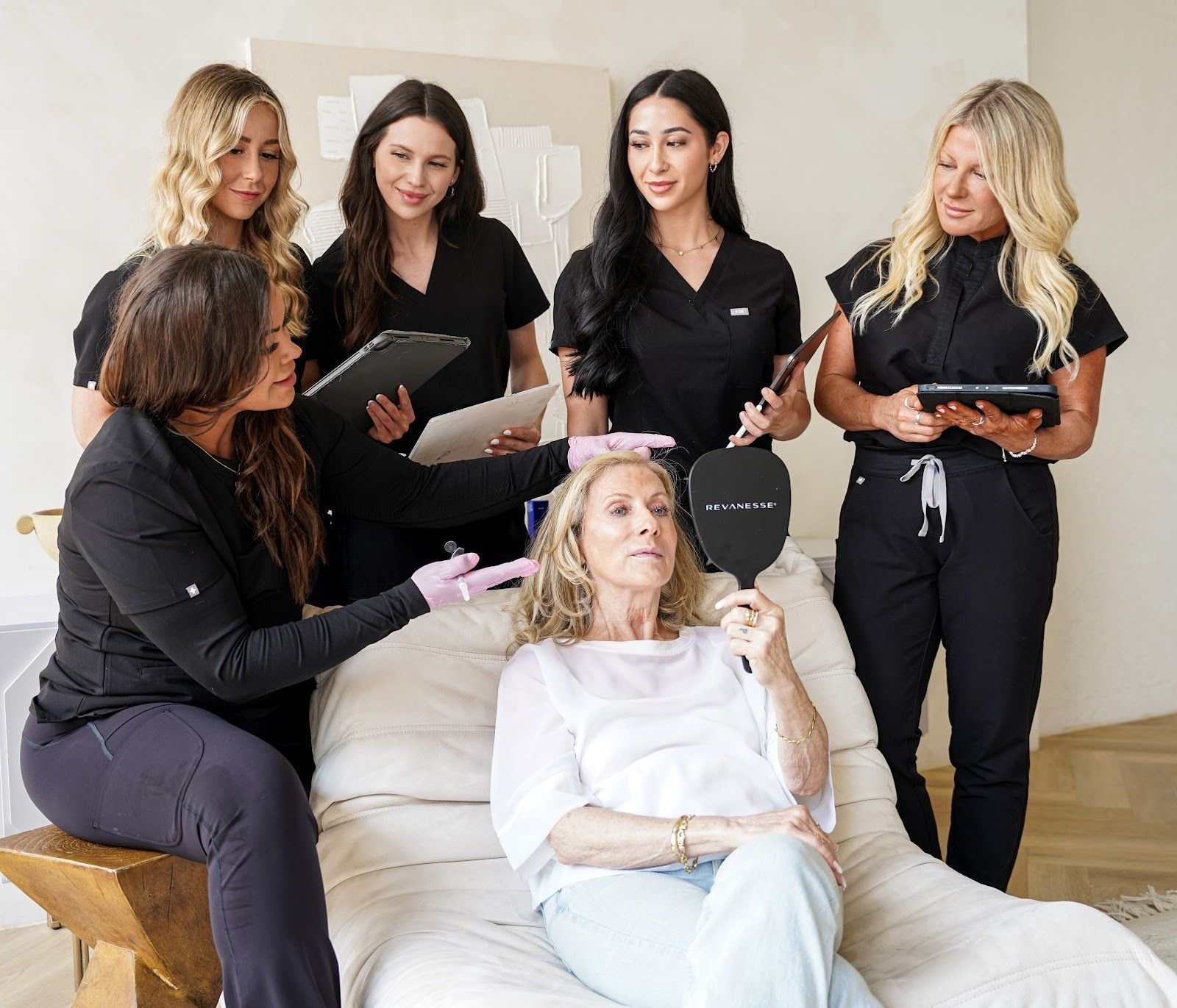 Five staff members in black uniforms consult with a woman holding a mirror in a bright, modern medical spa setting.
