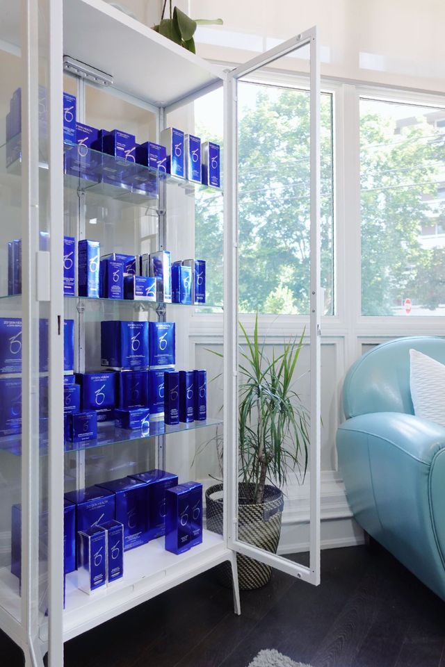 White cabinet with glass doors displaying blue skincare products, near a window and blue sofa.