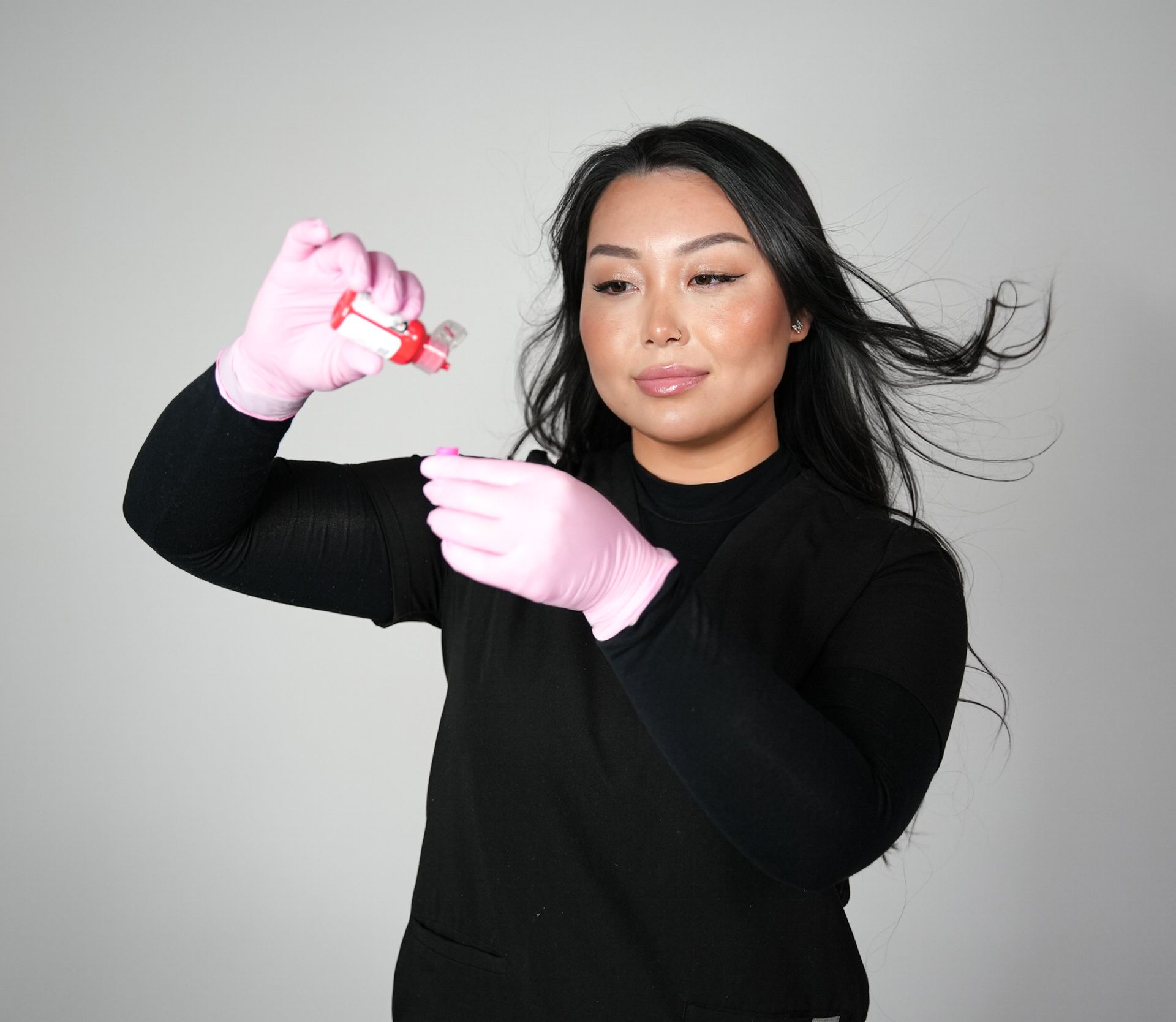 Woman in pink gloves holding a small bottle and tool, likely for beauty application. Black shirt, light background.