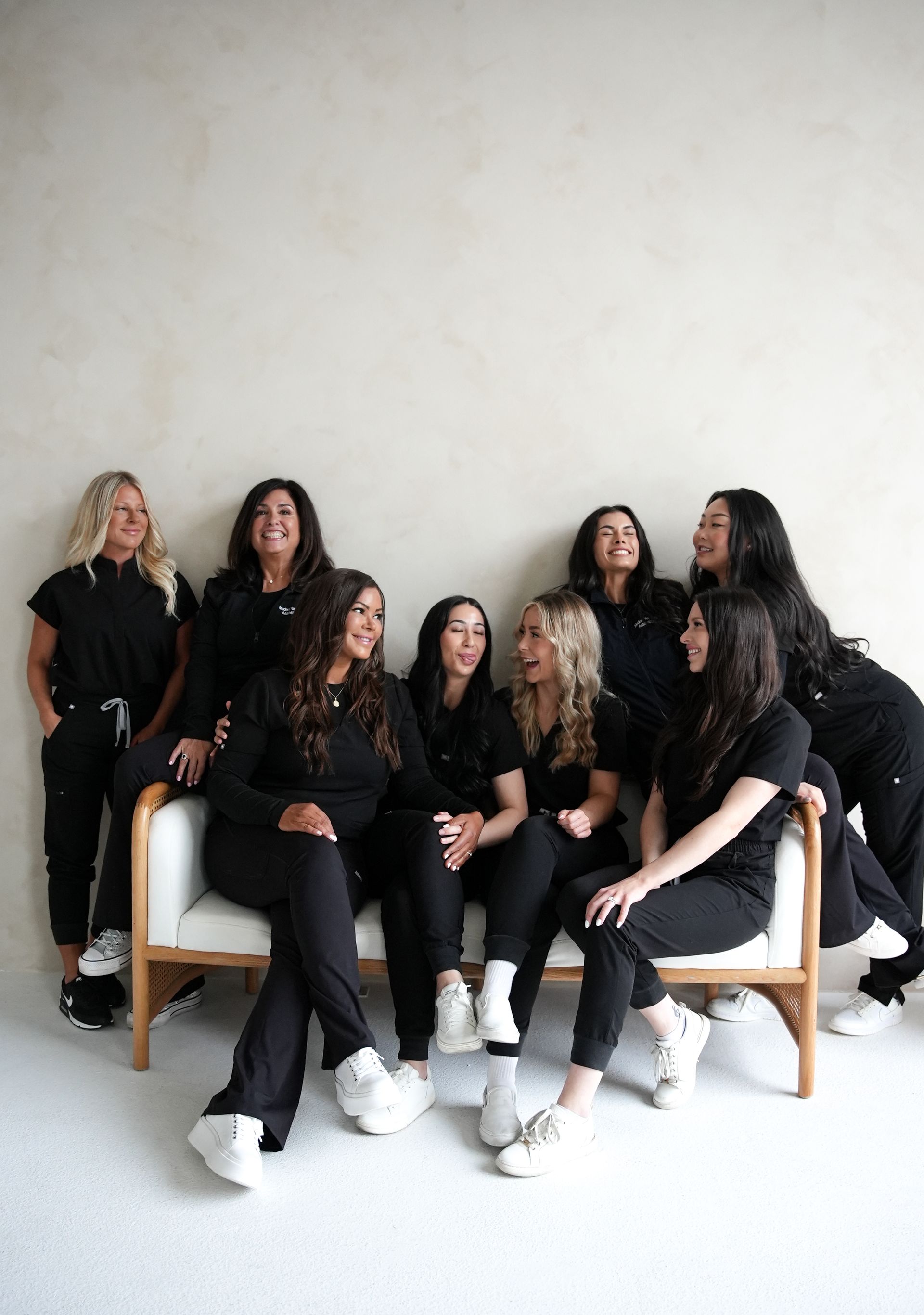 Nine women in black scrubs pose on a white couch, smiling, against a light background.
