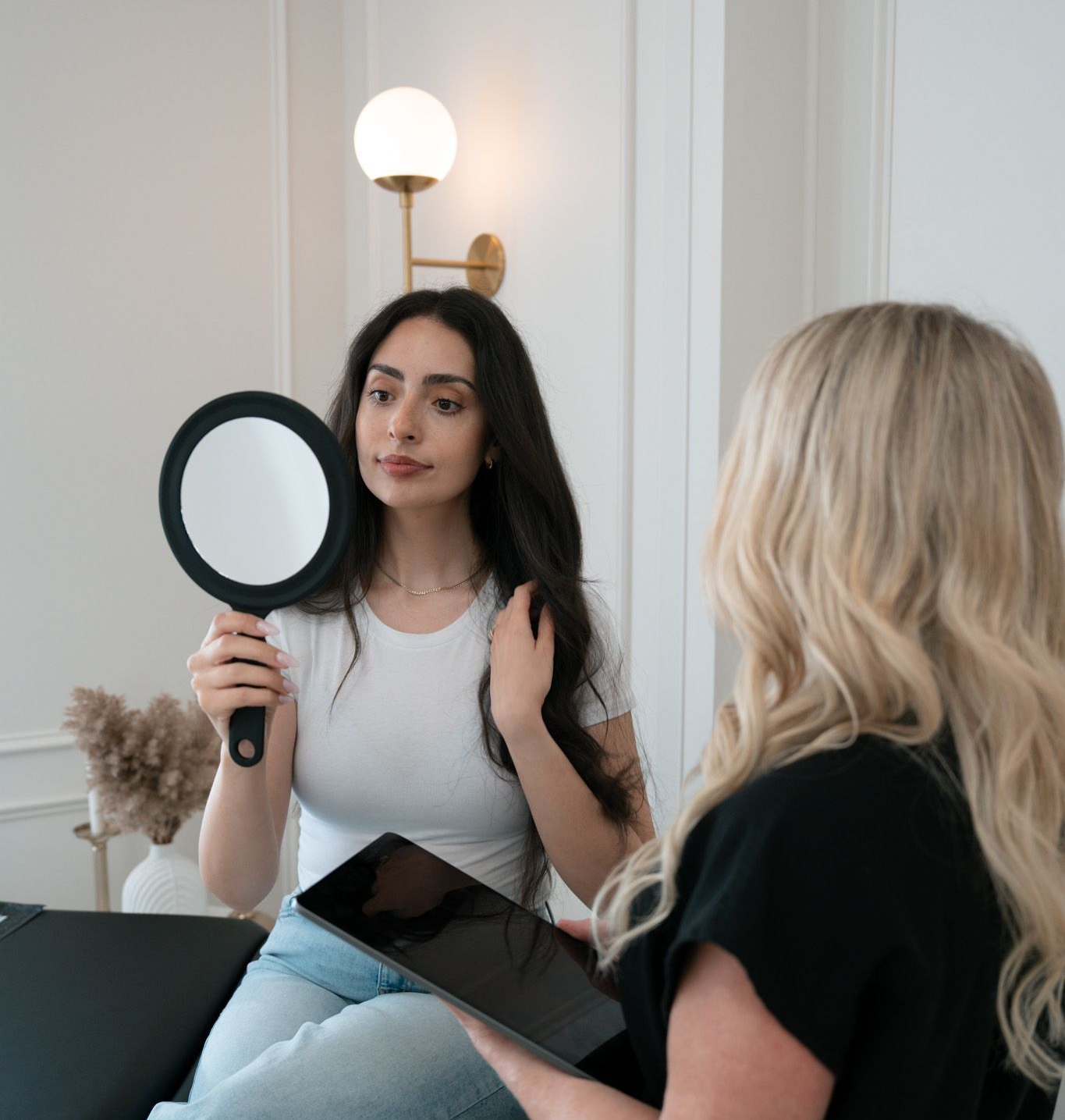 Two ladies one holding a mirror looking at her self with dark hair and touching her hair another with blonde hair standing and observing