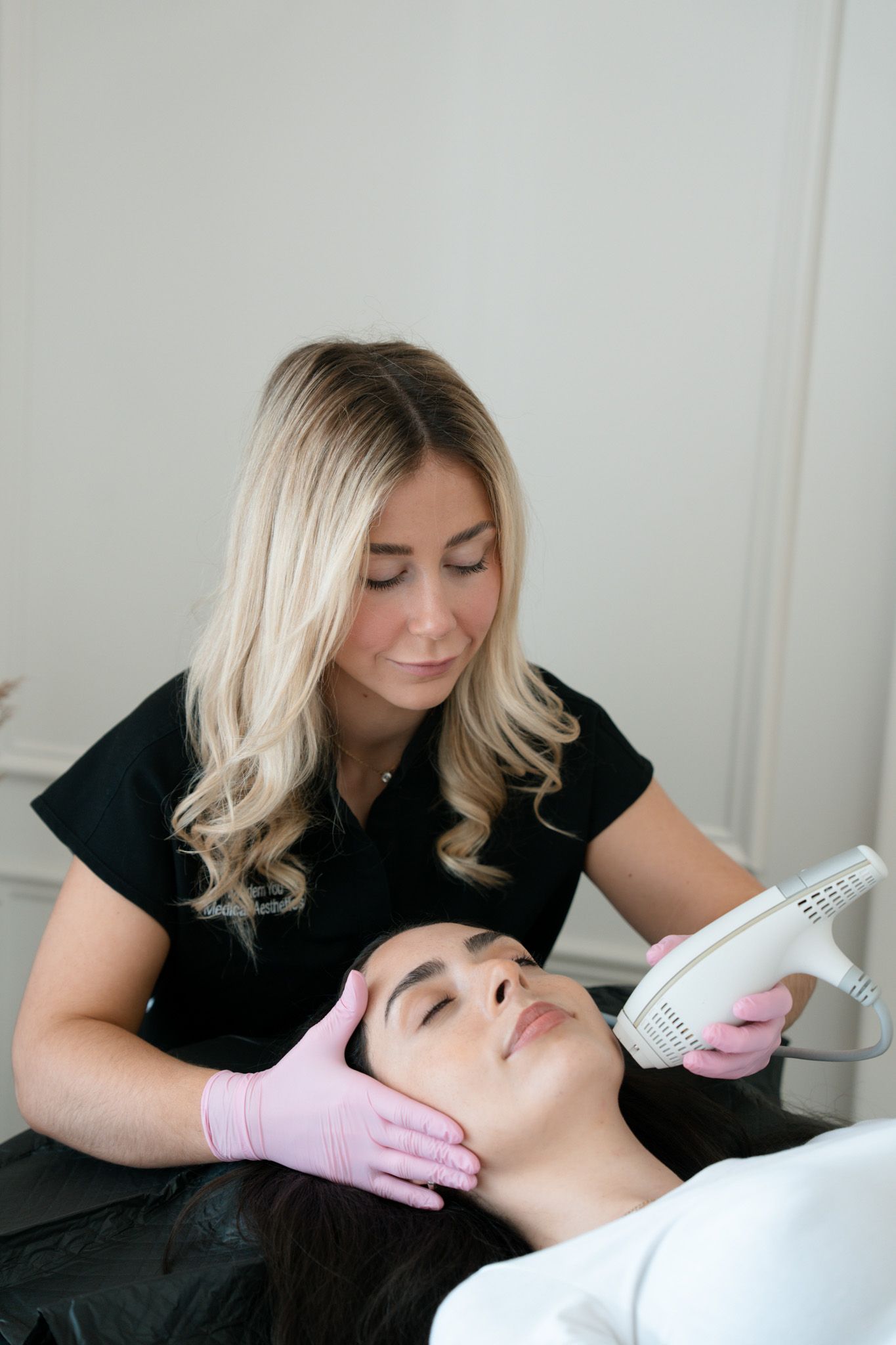 Woman receiving a facial treatment. Aesthetician in pink gloves holds device to client's face in a white-walled room.