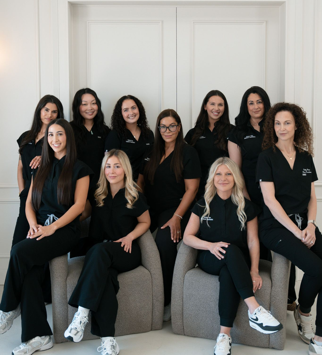 Eight women in white tops and jeans, holding hands, smiling, in a studio.