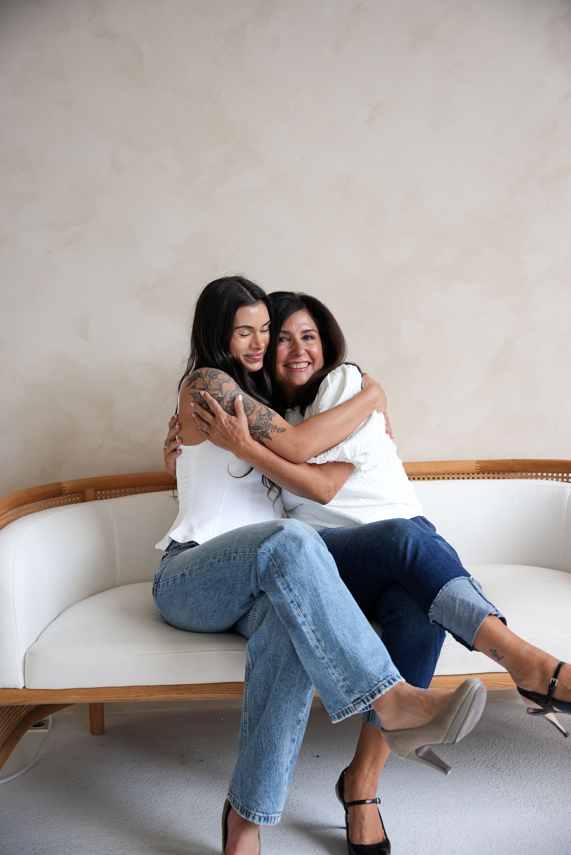 Two women embrace on a white sofa. One has tattoos, both wear jeans and white tops, smiling. Neutral-toned background.