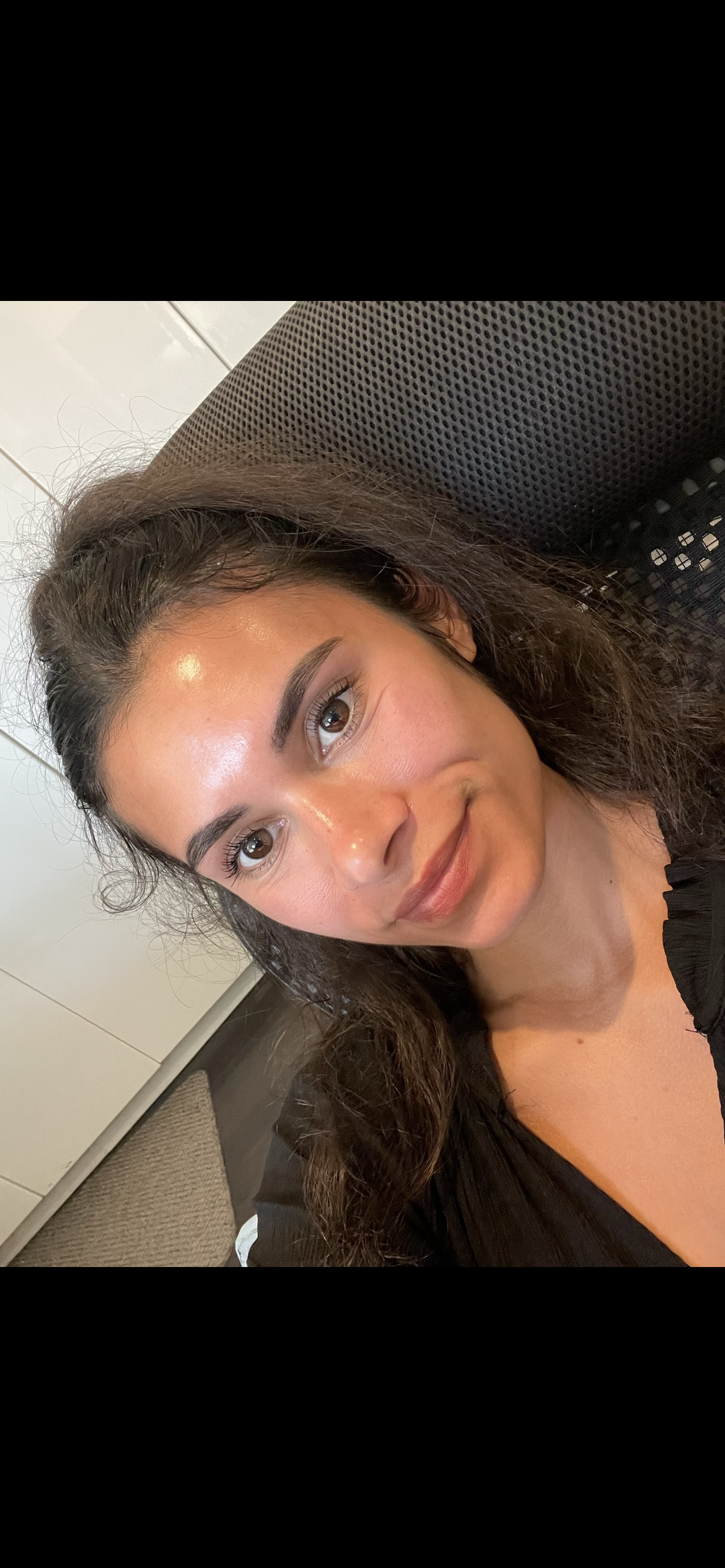 Woman with curly hair smiles, facing the camera. She is in a chair with a patterned back.