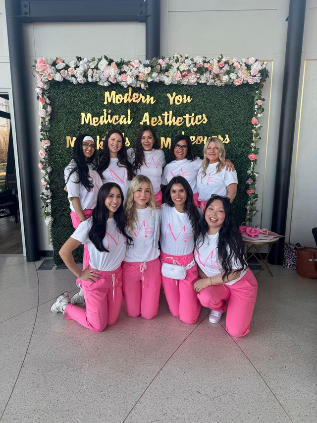 Group posing in front of a floral backdrop with gold “Modern Yet Medieval Aesthetics” lettering, wearing pink and white outfits