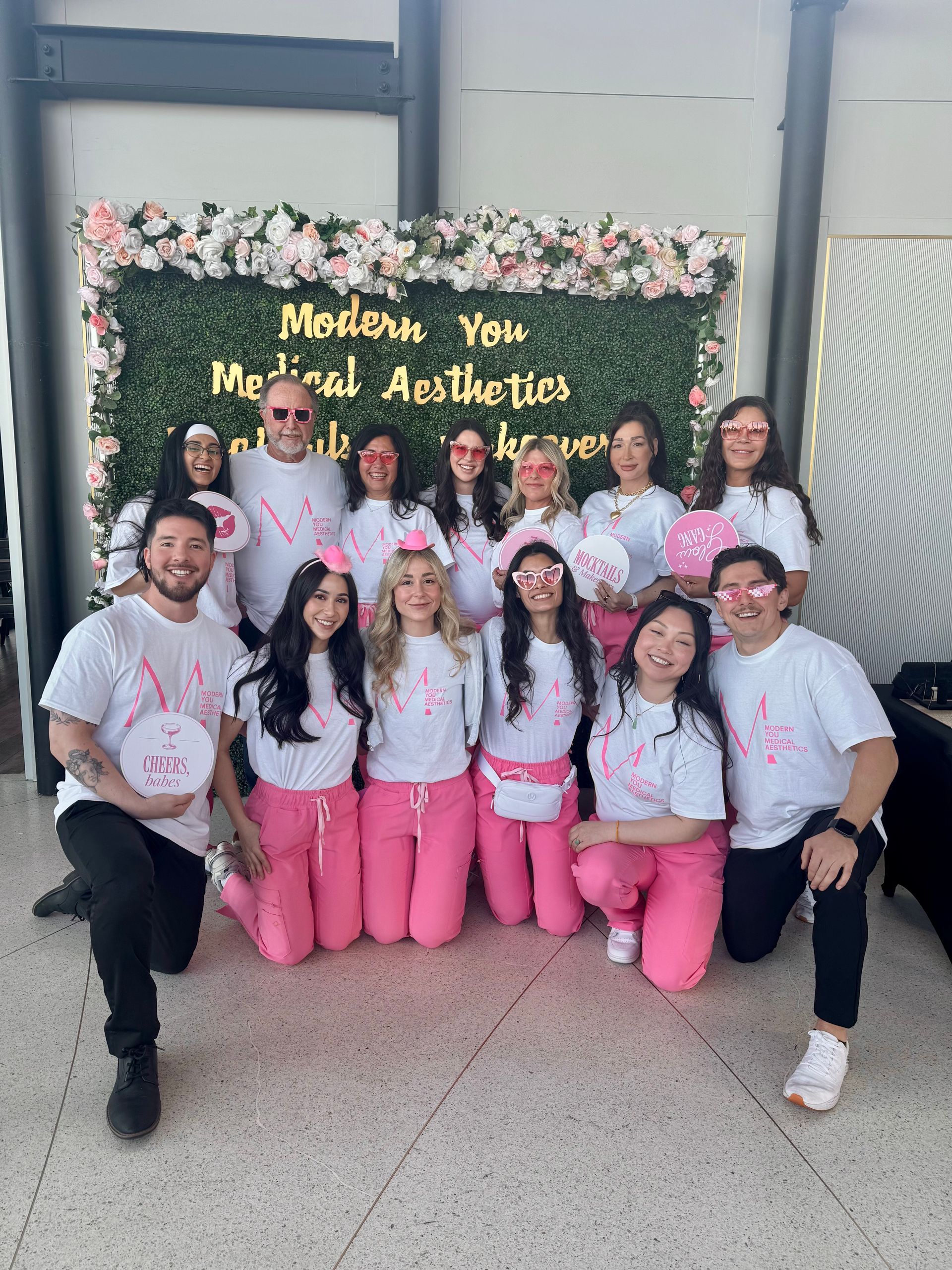 Group of people posing in pink and white outfits before a floral backdrop with gold anniversary lettering.