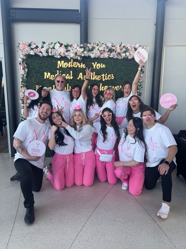Group posing in front of a floral backdrop, wearing white tops and pink pants, smiling at an event banner