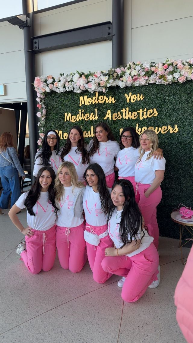 Group posing in pink pants before a flower wall with “Welcome New Medical Assistants” sign